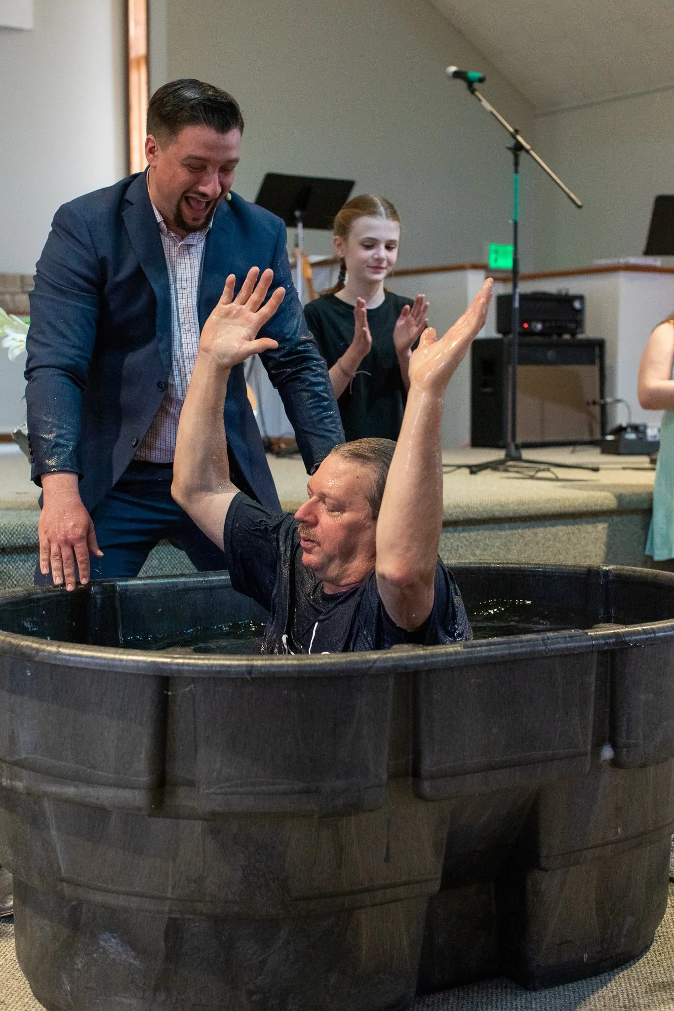 A man is in a baptismal pool with water, being baptized by another man. A woman and a girl are standing nearby, clapping and smiling.