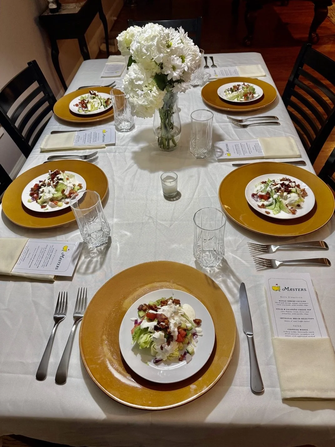 A formal dining table set with five plates of salad, clear glasses, white napkins, and a large bouquet of white flowers as a centerpiece.