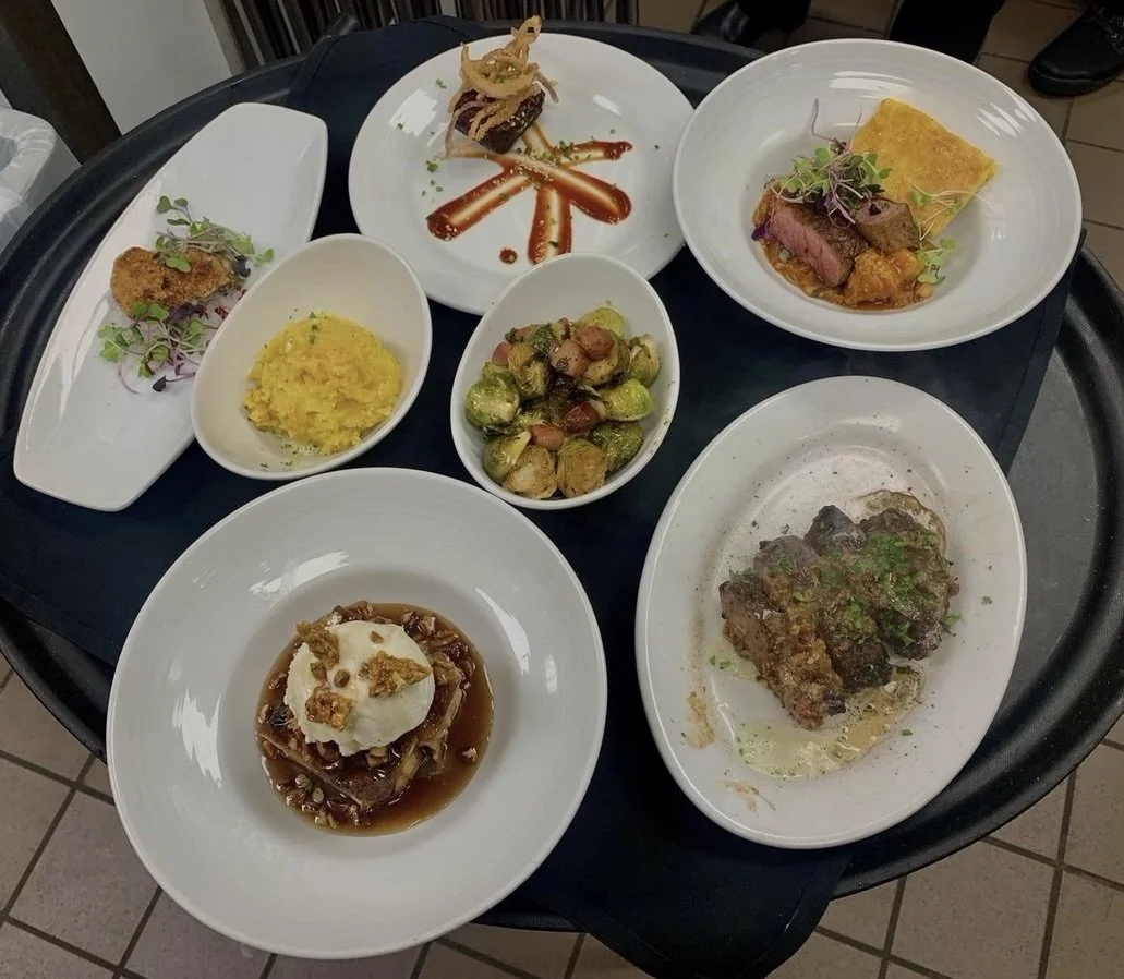 A black tray holding seven plates of assorted gourmet dishes, including fries, roasted vegetables, steak, meatloaf, and desserts.