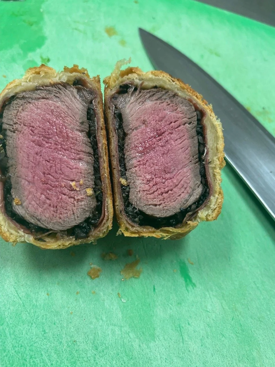 A halved beef Wellington on a green cutting board with a large kitchen knife nearby.