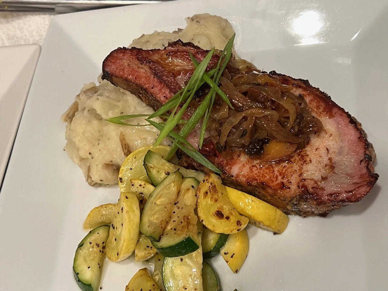 Grilled pork chop topped with caramelized onions, served with garlic mashed potatoes, roasted yellow squash, and garnished with green onions on a white plate.
