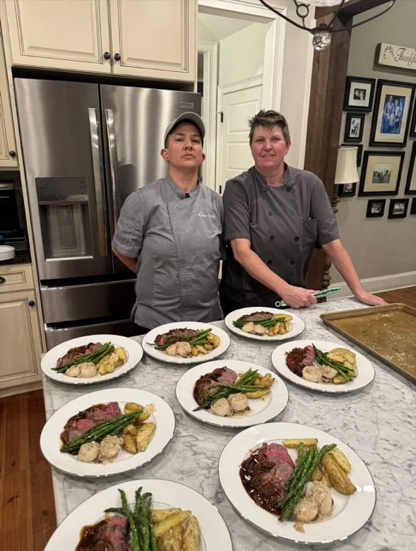 Two women in chef coats standing behind a kitchen counter with seven plates of cooked meat, green beans, potatoes, and gravy.