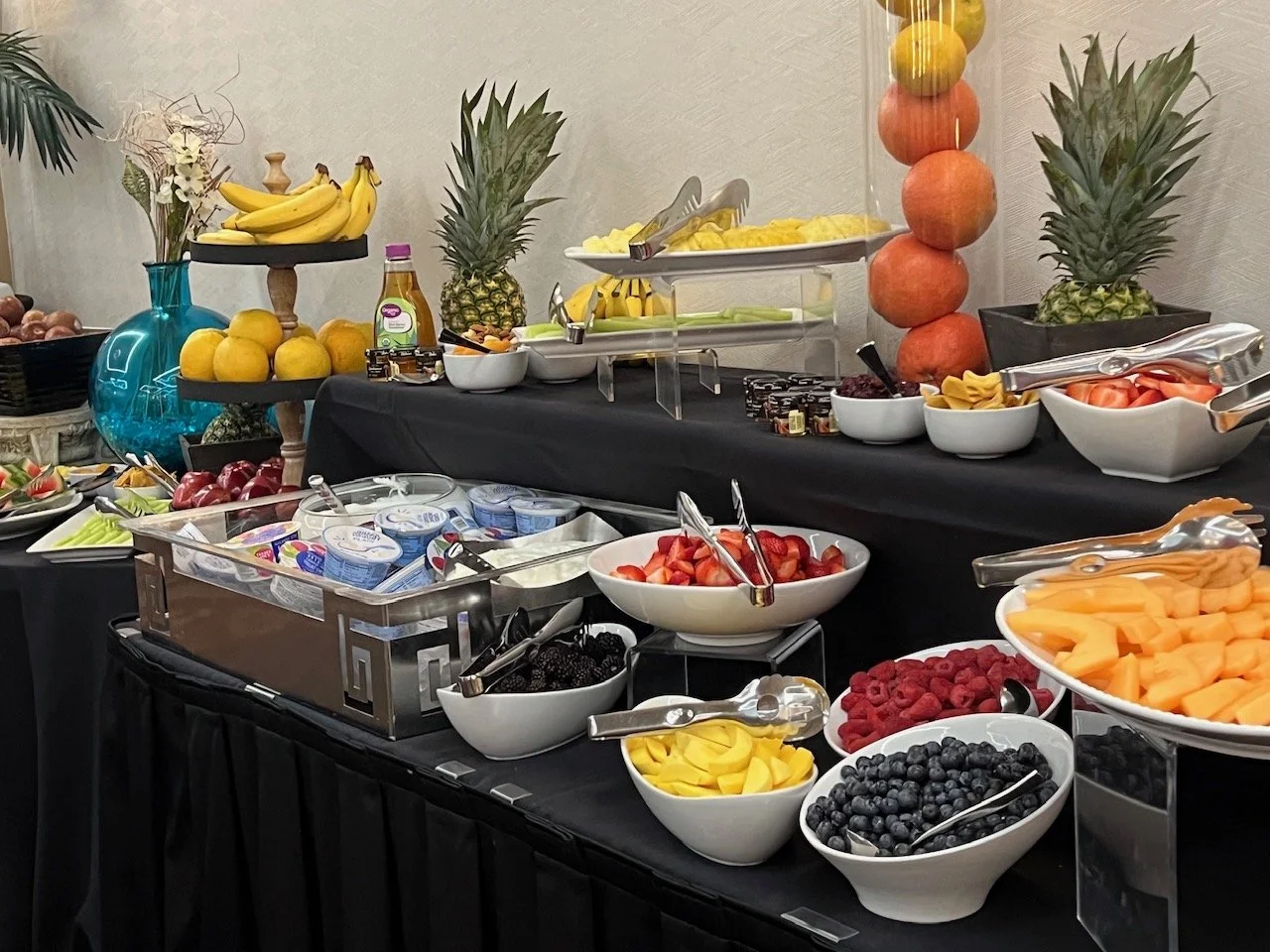 A buffet table filled with various fresh fruits including blueberries, strawberries, raspberries, blackberries, cubes of cantaloupe, honeydew, watermelon, sliced strawberries, raspberries, and blackberries, with pineapple and bananas visible in the background. The setup includes bowls, tongs, and utensils, with decorative pineapples and oranges.