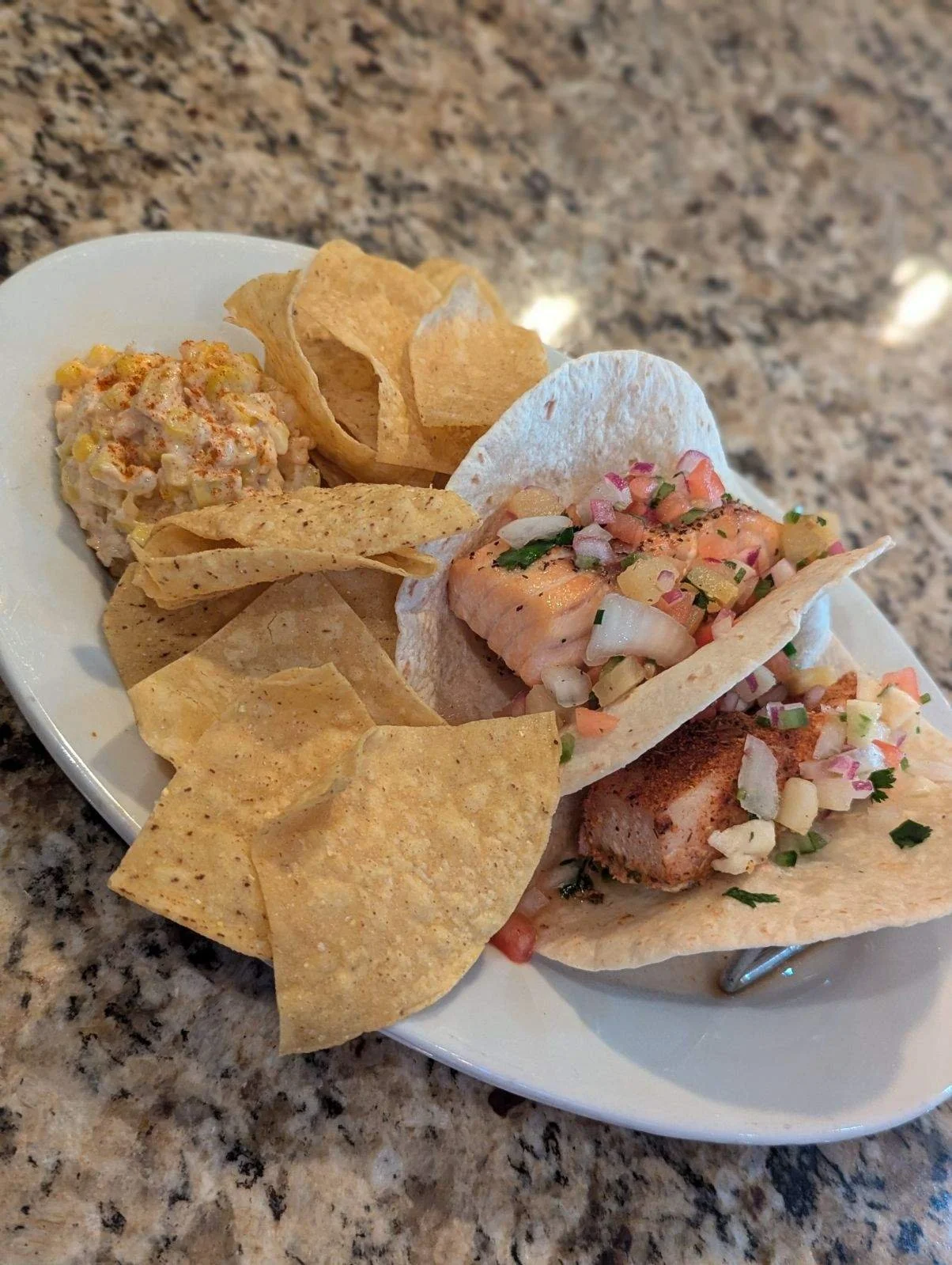 A plate with chips, a scoop of Mexican street corn salad, and two fish tacos topped with chopped onions, cilantro, and salsa, on a granite countertop.