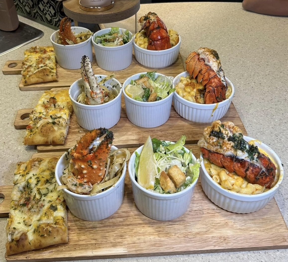 Arrangement of lobster dishes and appetizers on a wooden serving board, including mini pizzas, lobster tails, and salads.