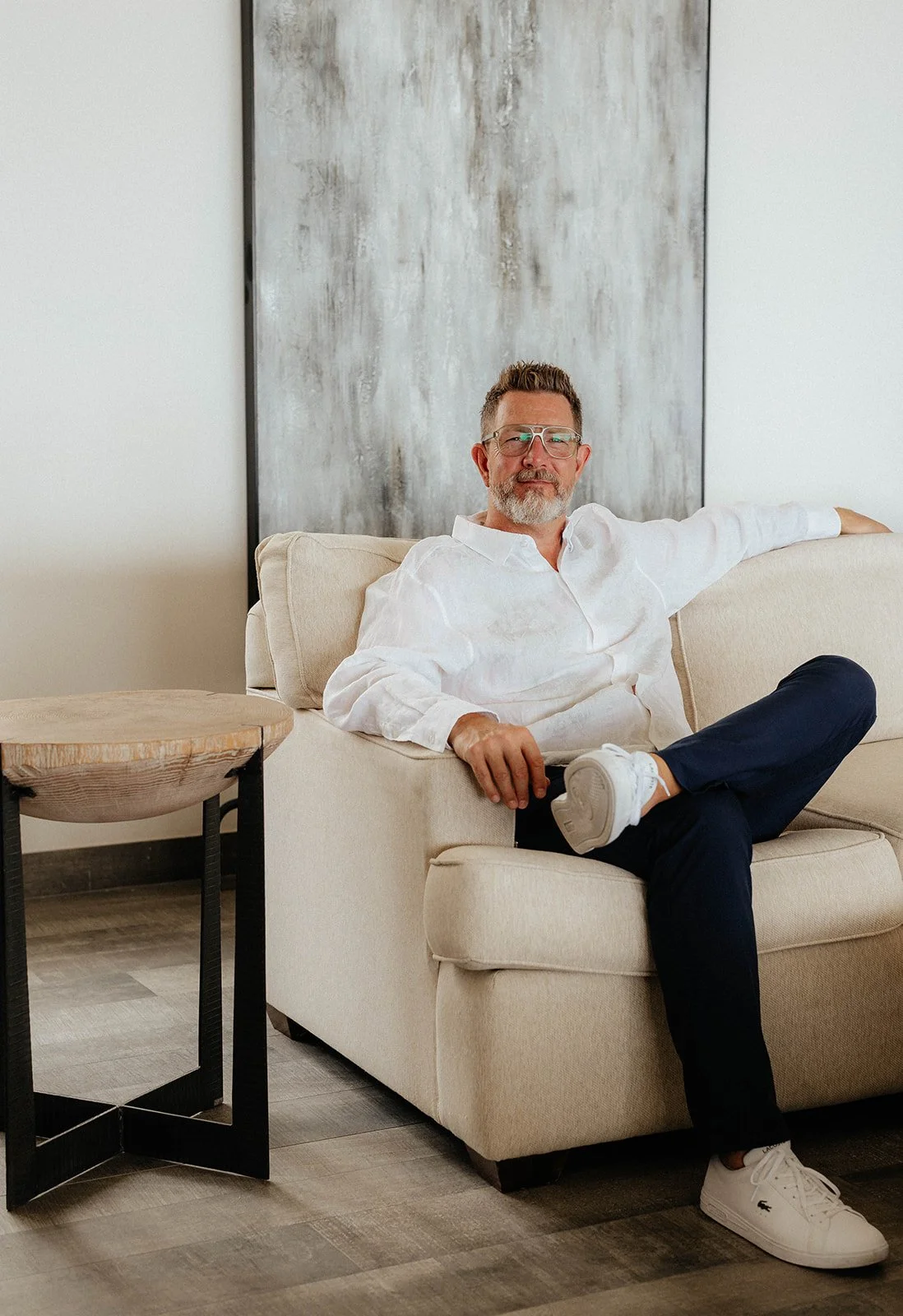 A man with glasses and a beard, wearing a white shirt and dark pants, sitting on a beige sofa with one arm resting on the backrest. He has one leg crossed over the other and is wearing white sneakers. There is a wooden side table with black legs next to the sofa. A large abstract painting is hung on the wall behind him in a minimalist living room.