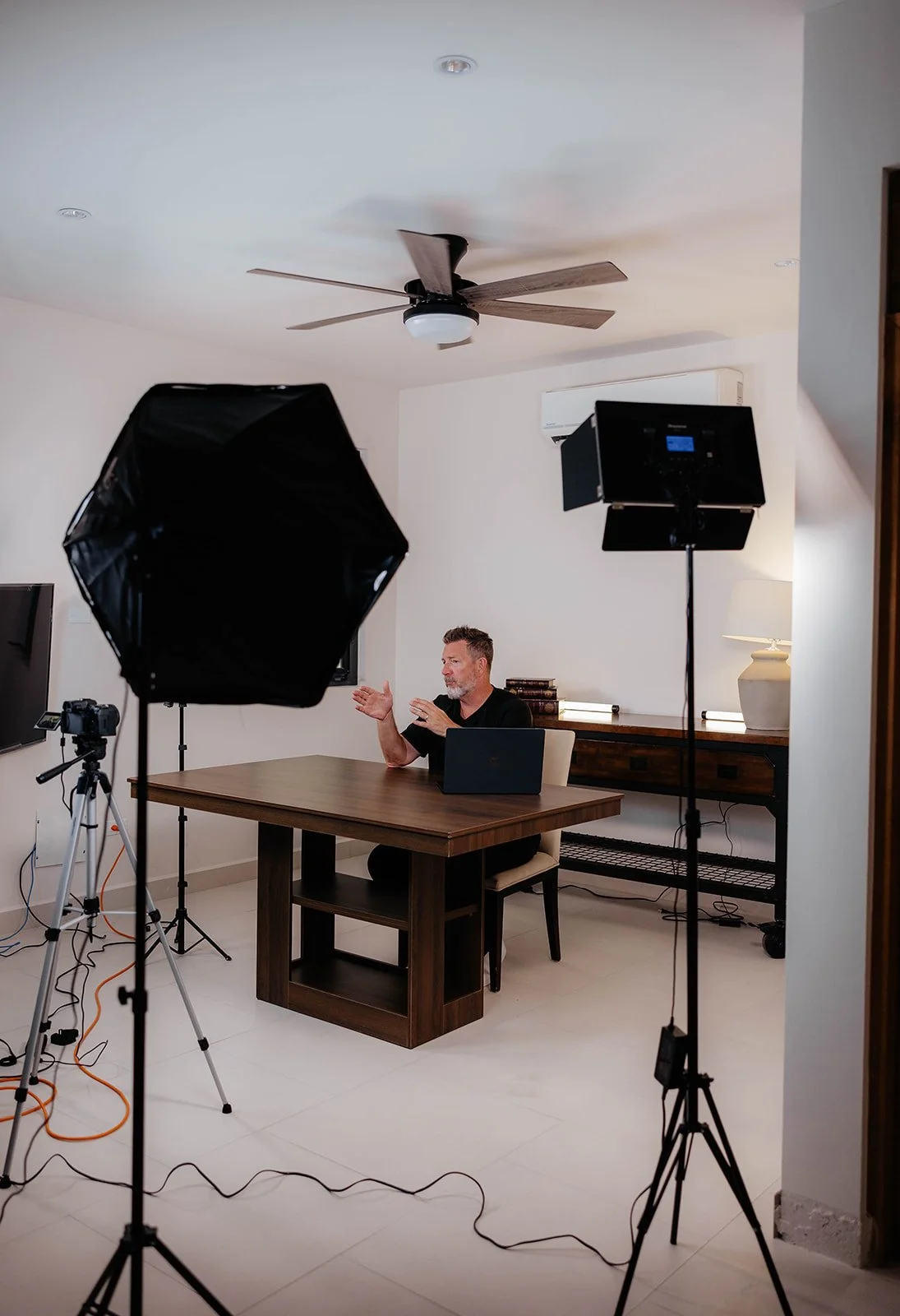 Man in black shirt sitting at a wooden table during a video recording or live stream setup, with lighting and camera equipment around him in a room with white walls and ceiling fan.