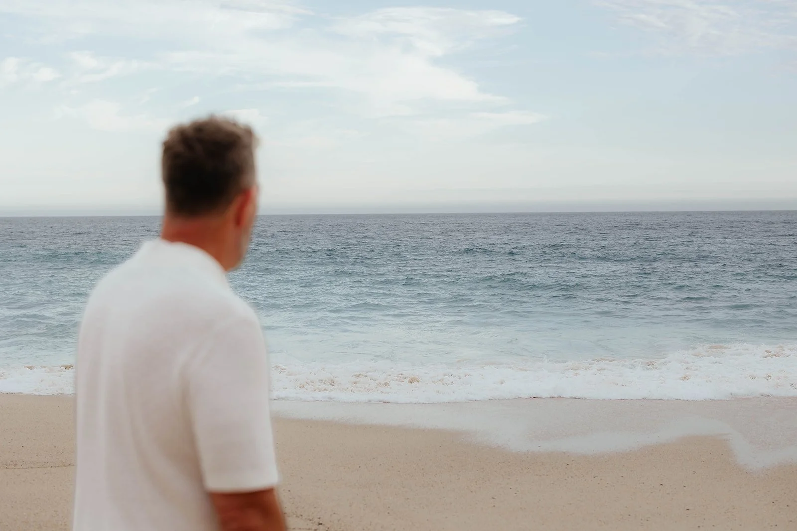 A man, Dr. Nathan Goodyear, with short hair wearing a white shirt, facing the ocean on a sandy beach during the daytime.