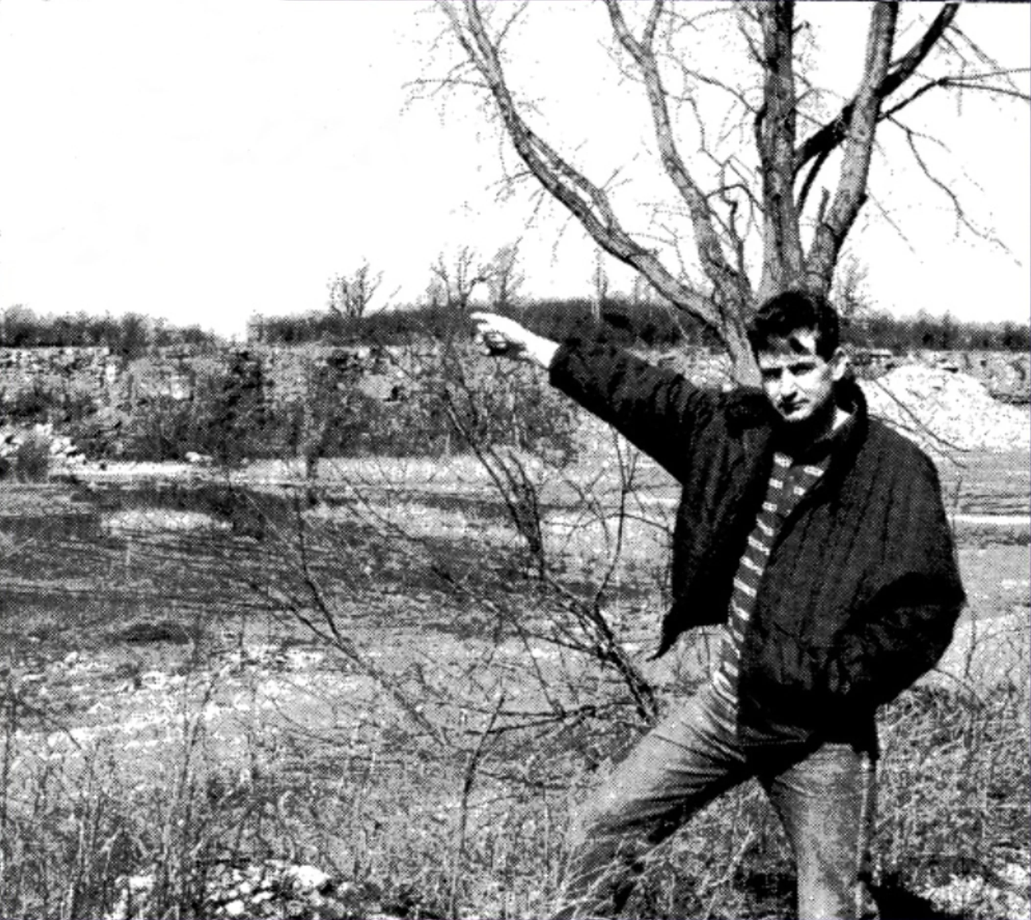 Pat McCarthy wearing a coat and jeanss leaning against a tree with outstretched arms in an outdoor rural setting pointing to where he saw the UFO.