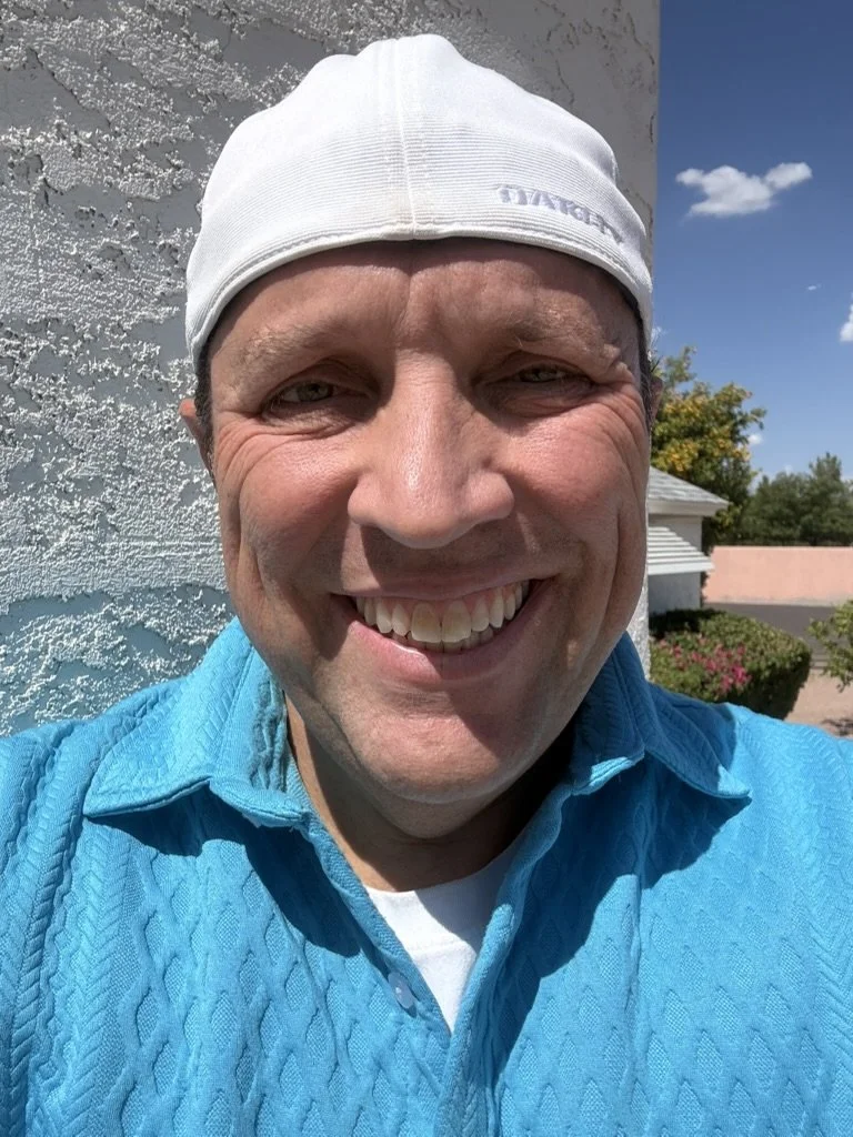 A man smiling outdoors wearing a white cap and a blue shirt, with a textured white wall behind him and a blue sky with some clouds.