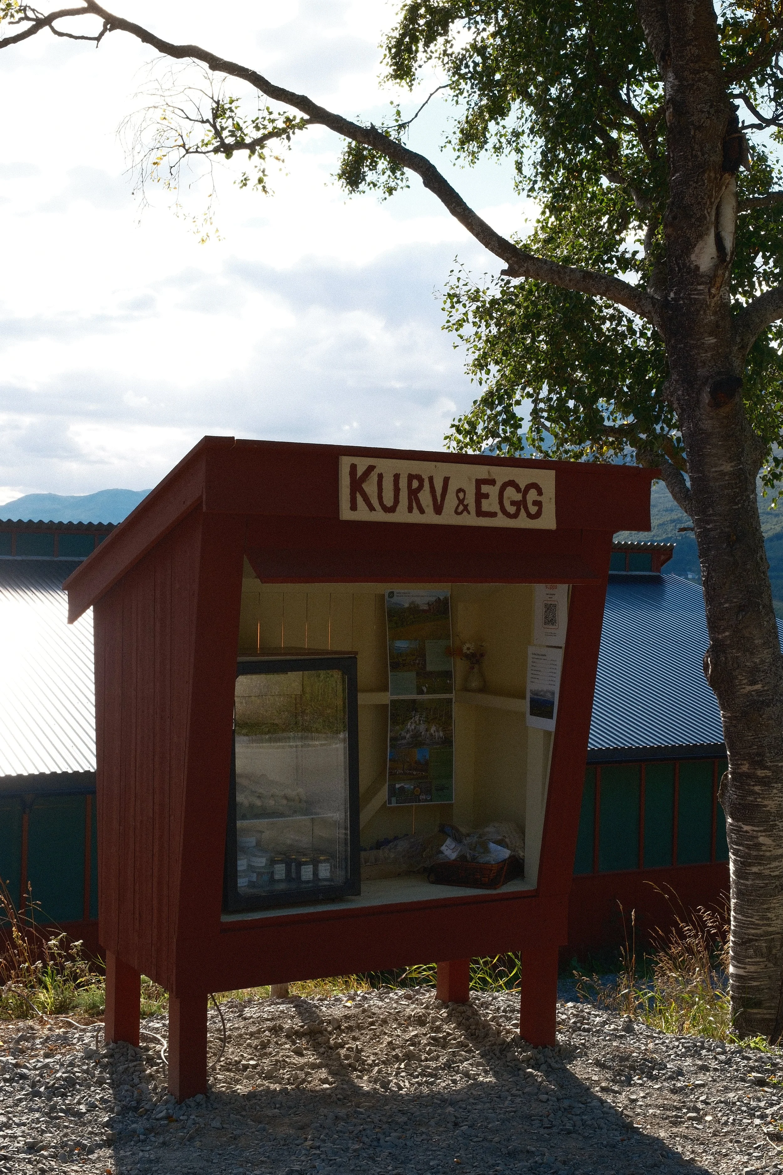 Rødt informationsskilt med teksten «KURV & EGG» foran en svartklistret bygning, med et tre til høyre og fjell i bakgrunnen under en overskyet himmel.
