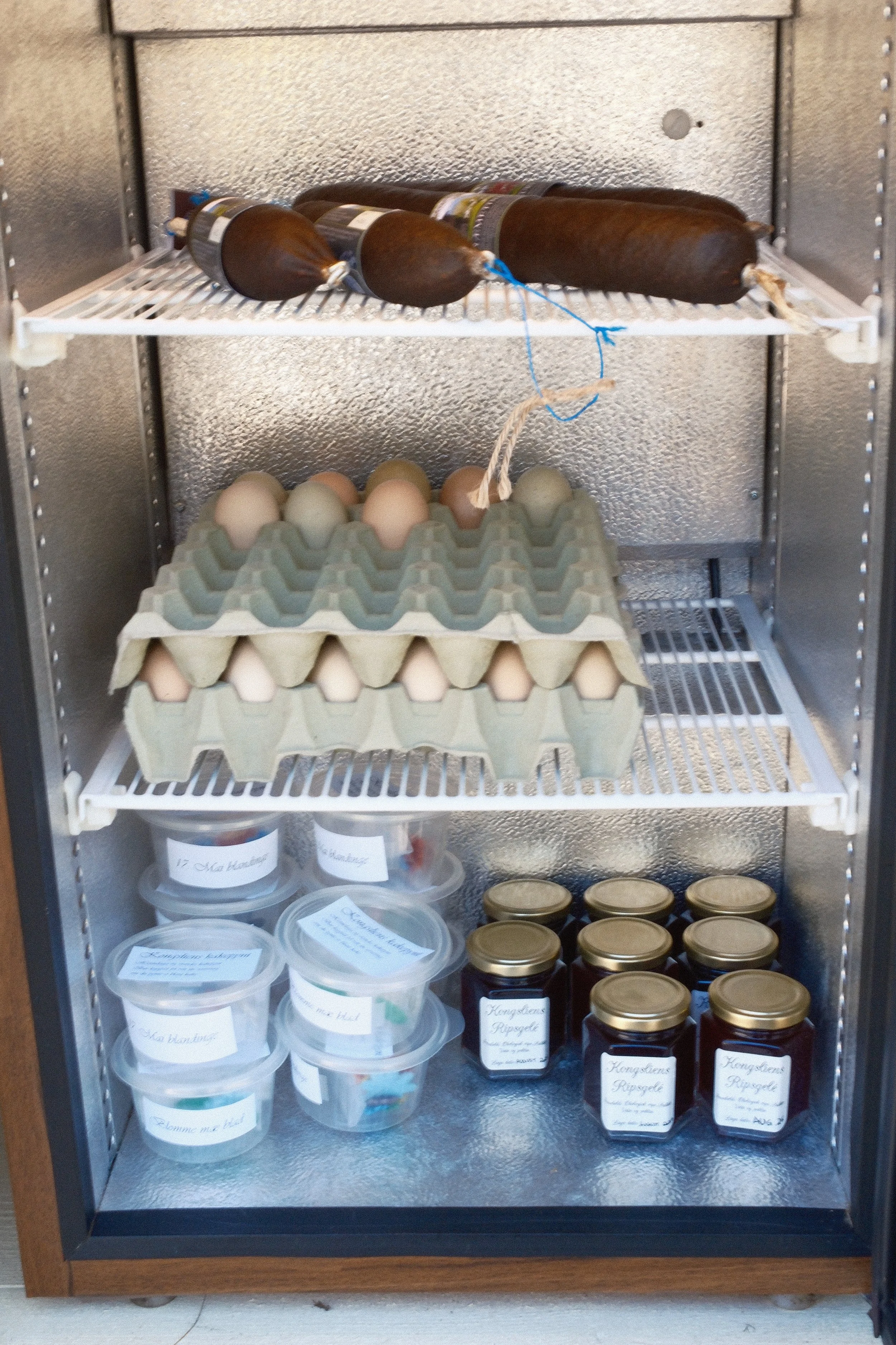Oppbevaringsskap med egg, viltpaté og syltetøy. Egg i pappesk, glass- og plastbeholdere med etiketter, og små glass med gulltopper.