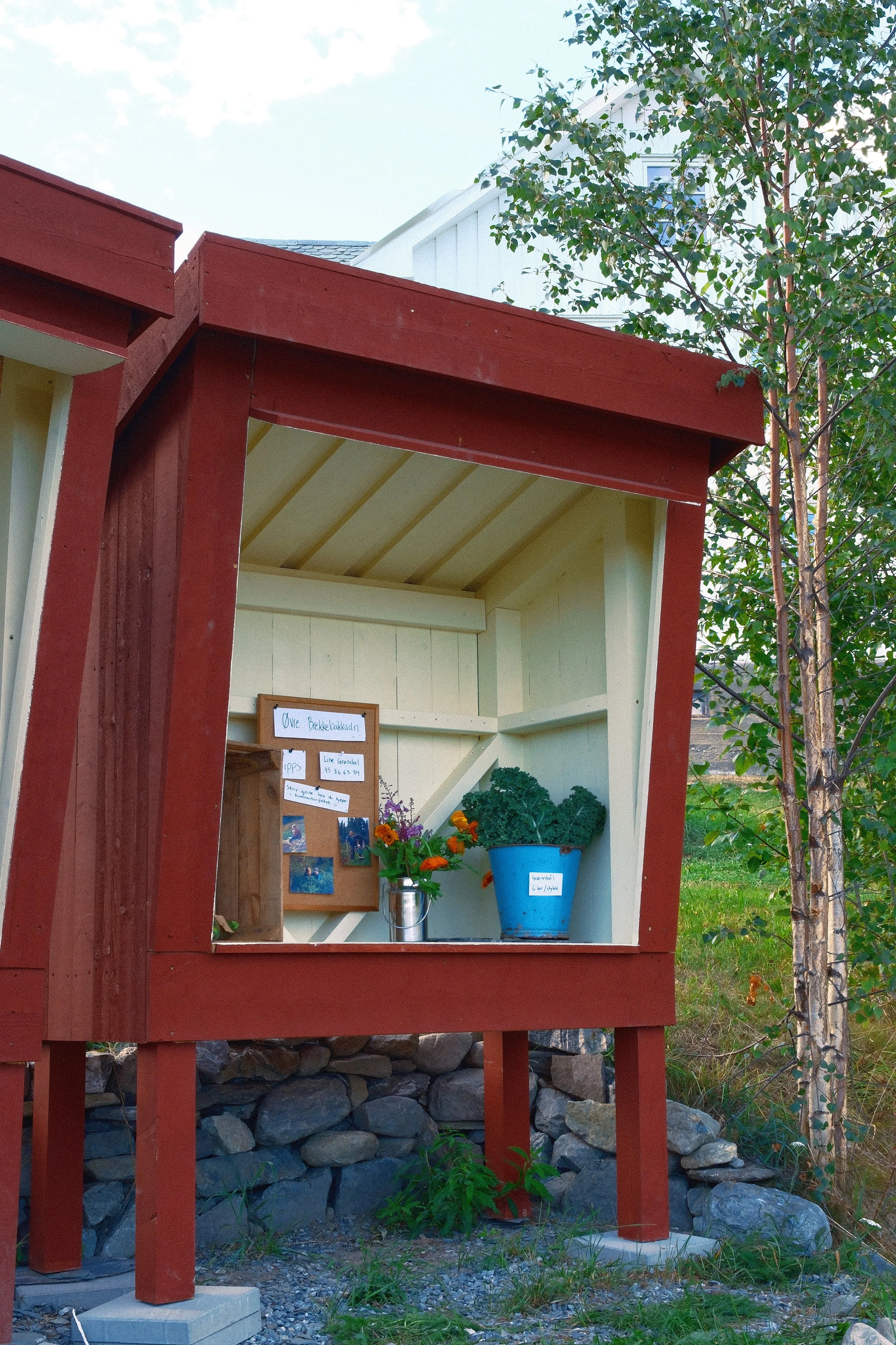 Et lite hus med rødt utvendig og hvit innside, plassert på en opparbeidet grunn med steiner og gress, omgitt av trær, med et oppslagstavle og blomster inne i utstillingen.