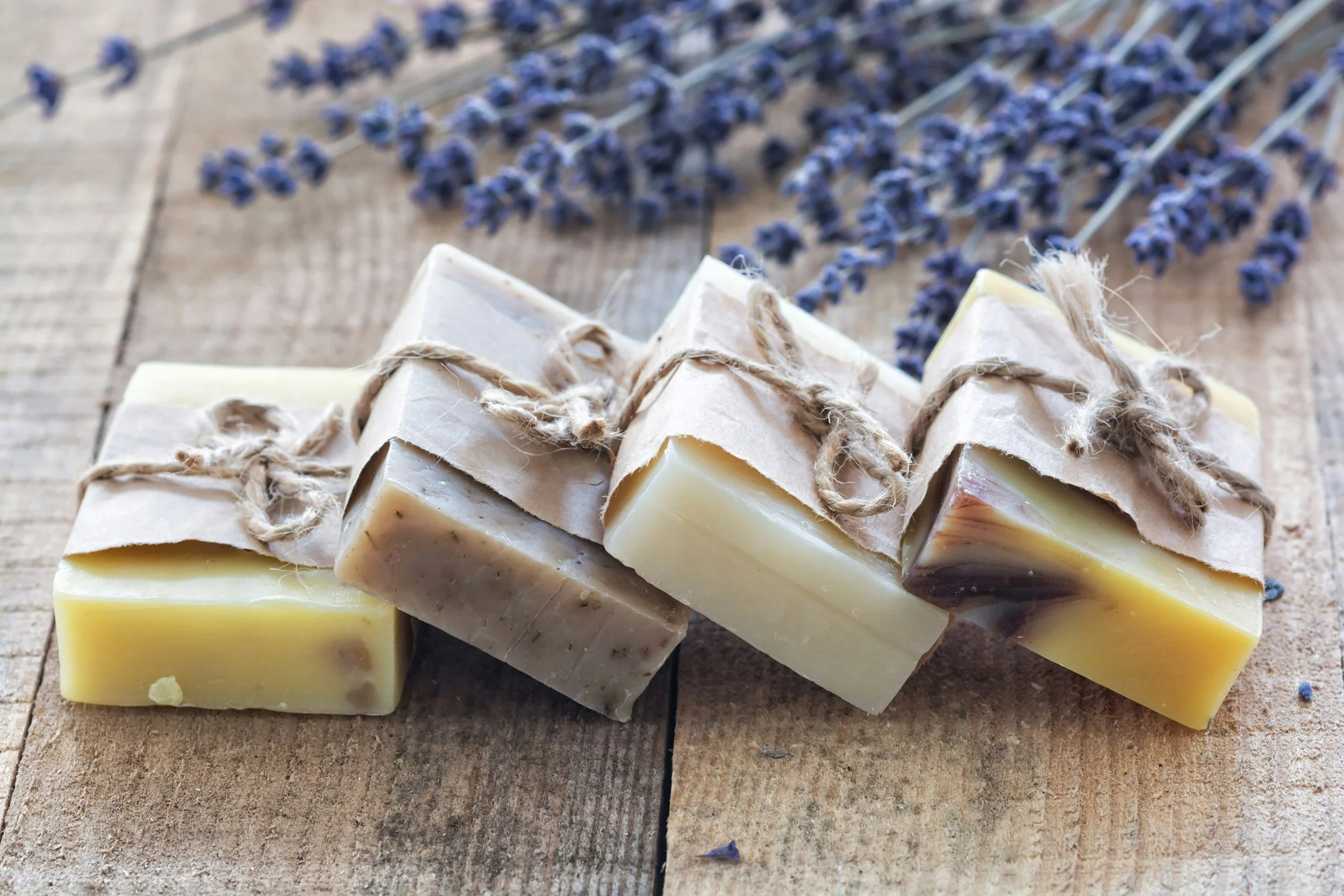 Utsnitt av flere håndlagde tova såper med naturlige ingredienser, lagt ut på en rustikk treflate, med noen lavendelblomster i bakgrunnen.