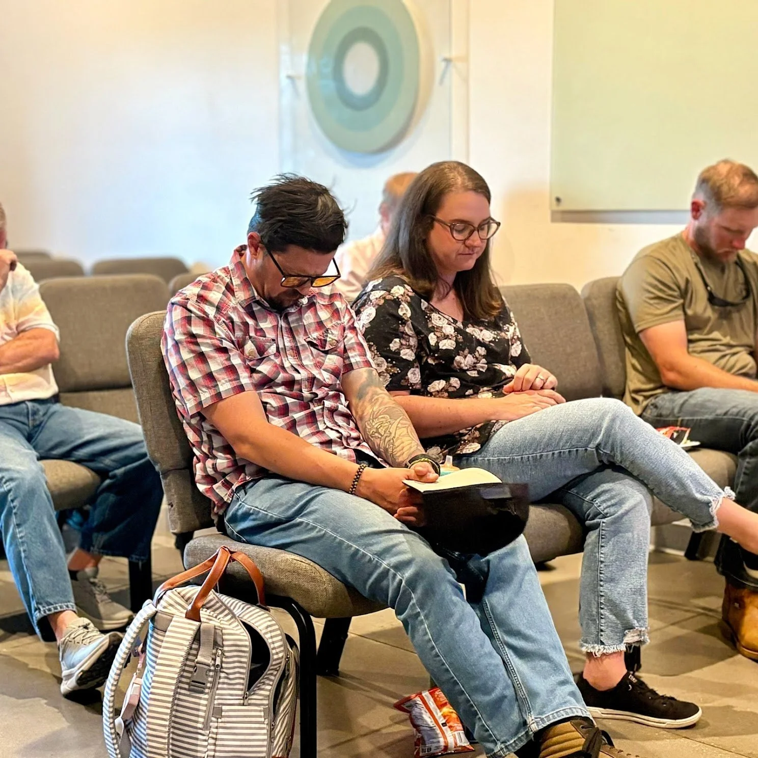 People seated on chairs listening attentively, some taking notes, in a conference or seminar room. The background features a circular target design on the wall.