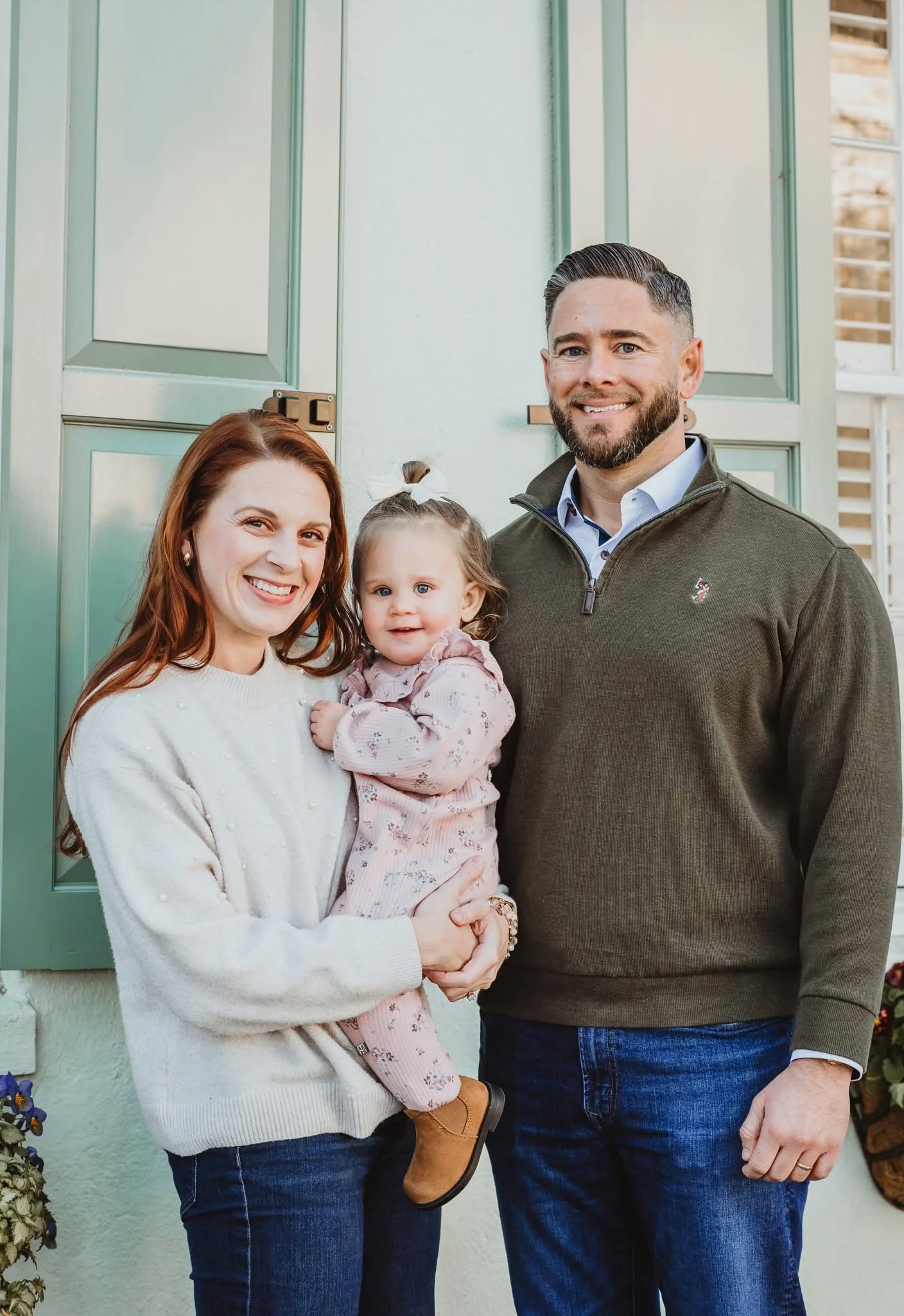 A smiling woman holding a young girl in front of a pastel green house door with a man standing beside them.