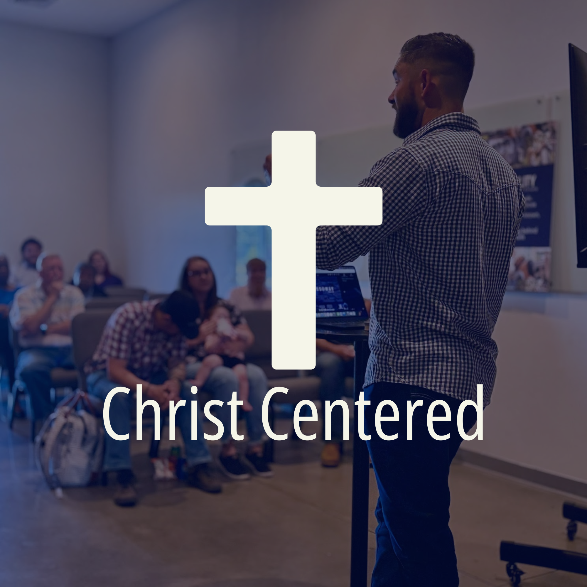 A man with short dark hair and a beard, wearing a checkered shirt, is speaking at a podium during a church event with a large congregation. The background shows seated attendees and a bulletin board.