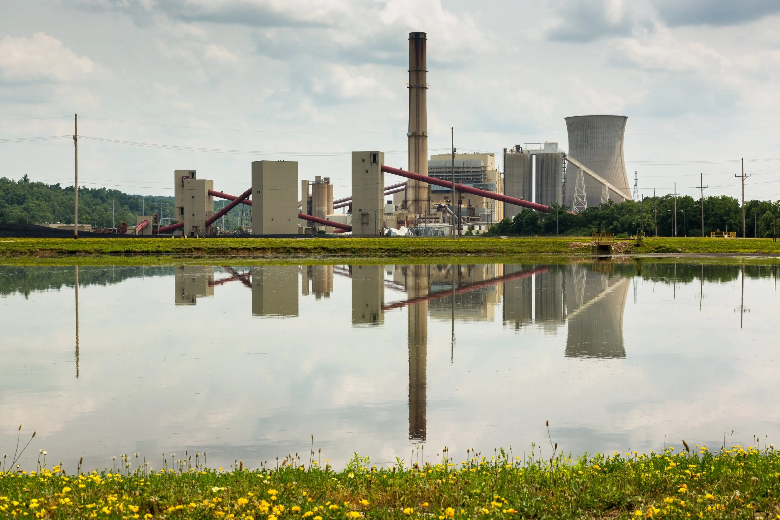 Industrial power plant with cooling tower and smokestack reflected in calm water, surrounded by green hills and yellow wildflowers.