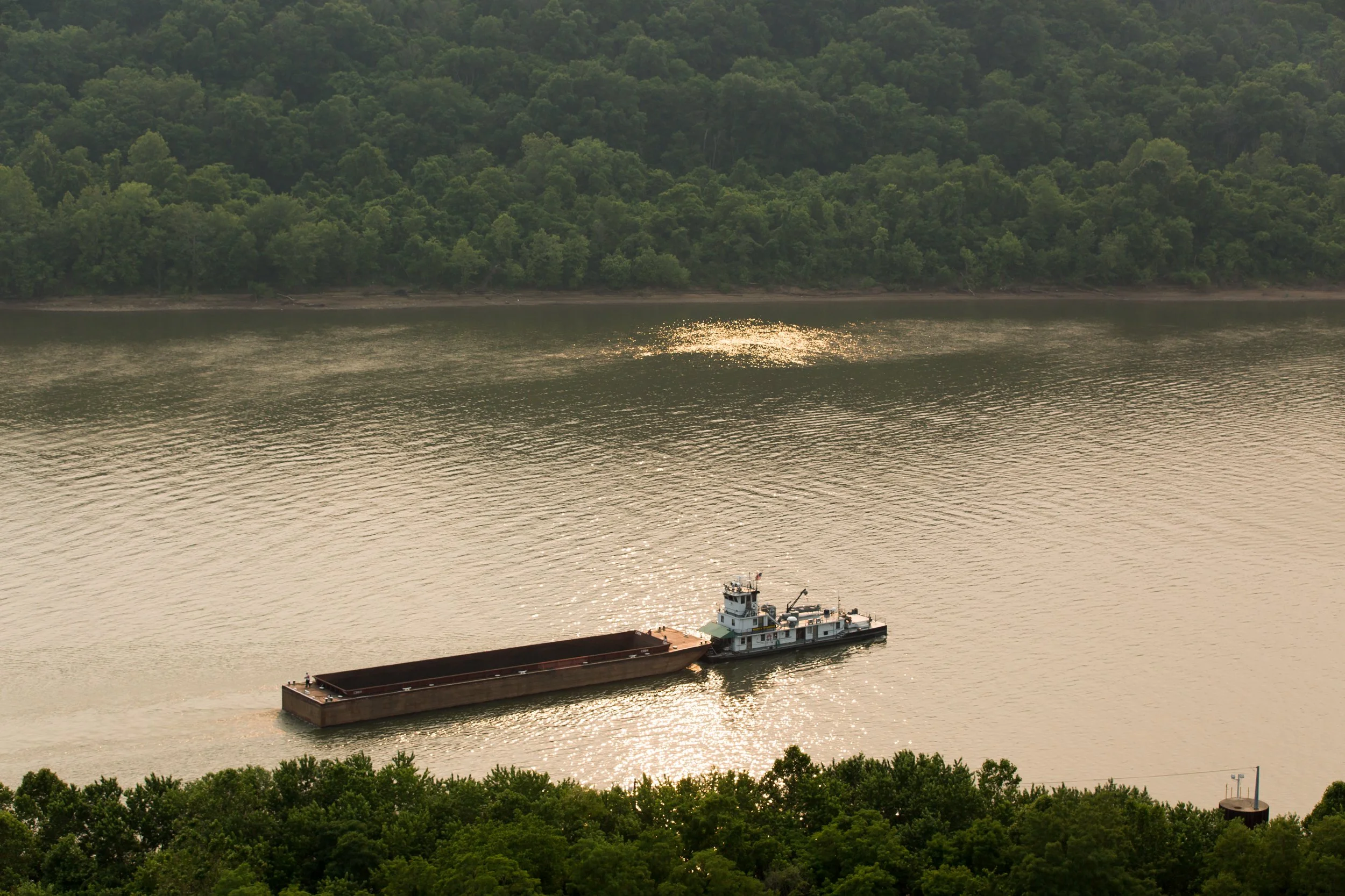 A cargo ship sailing on a river, with a forested hillside in the background and the sunlight reflecting on the water.