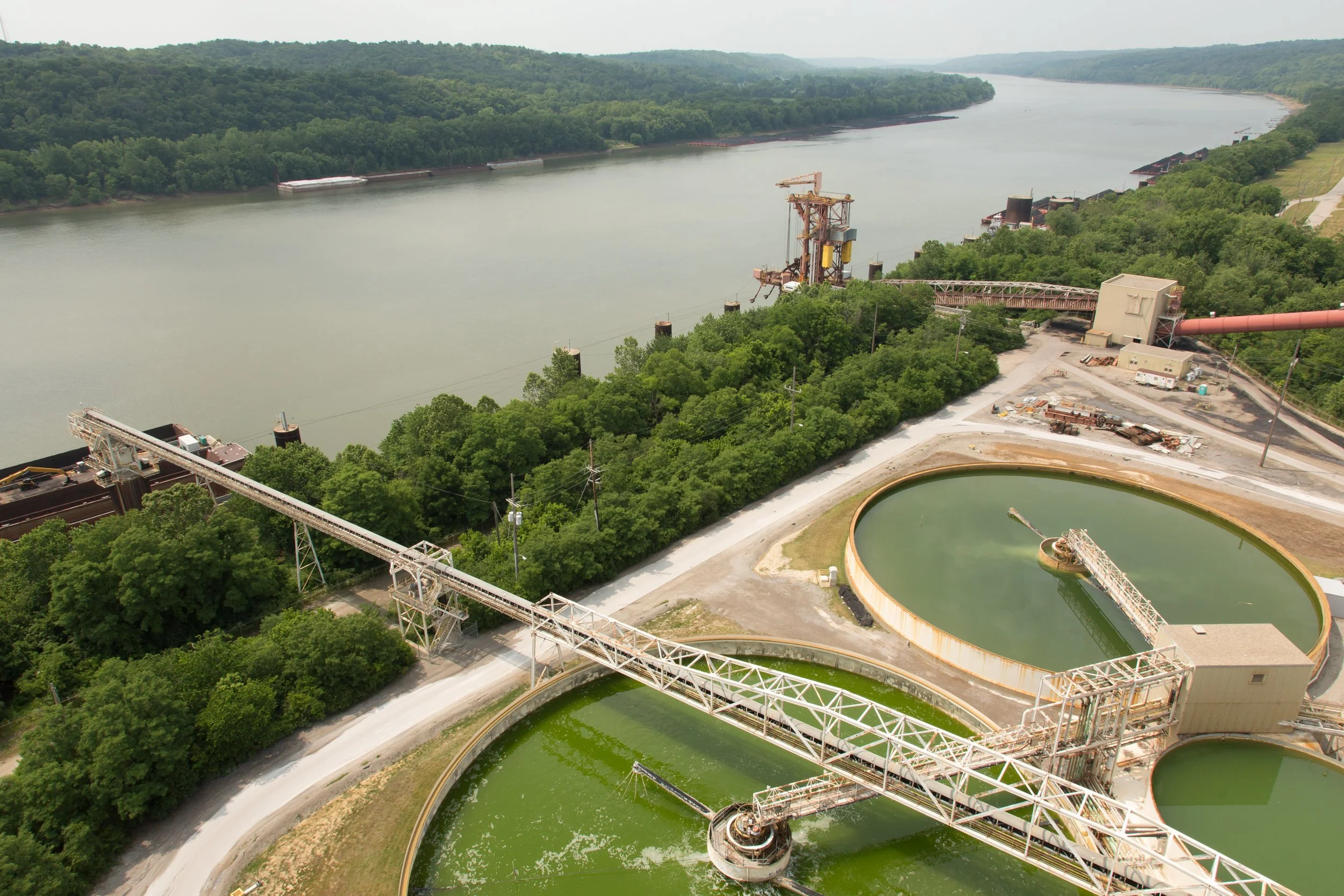 An aerial view of a water treatment plant with circular sedimentation tanks and a long conveyor system, surrounded by dense green trees and a river in the background.