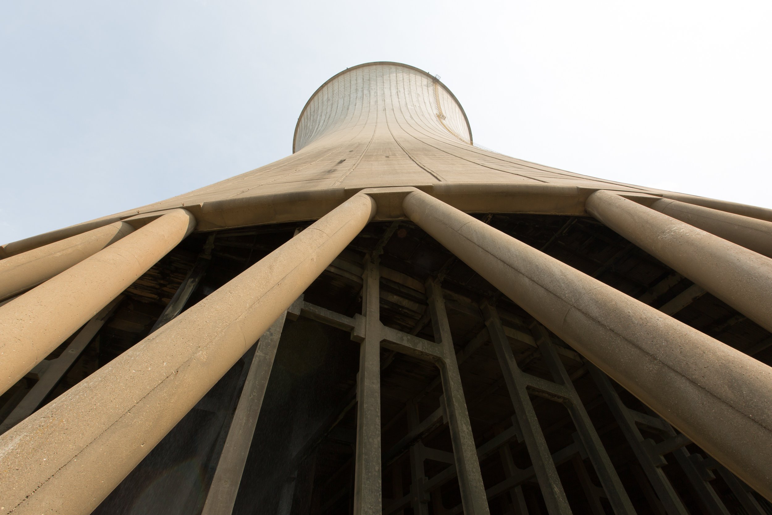 Low-angle view of a cooling tower with beige concrete support columns at the base, against a pale sky.