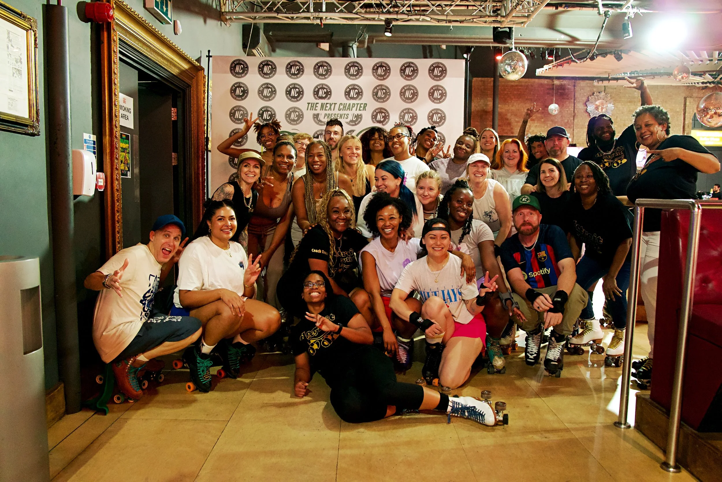 A large group of diverse people posing in a roller skating rink, smiling and making peace signs, with some wearing roller skates and casual attire, and a backdrop with logos and the text 'The Next Chapter Presents'.