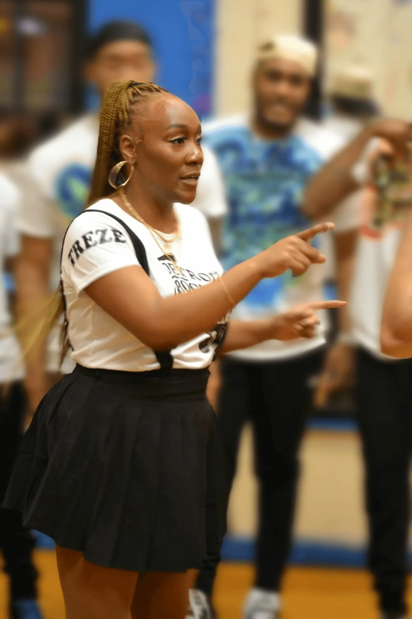 A woman with braided hair, hoop earrings, and gold jewelry pointing and talking in a group setting, with other blurred women in the background.