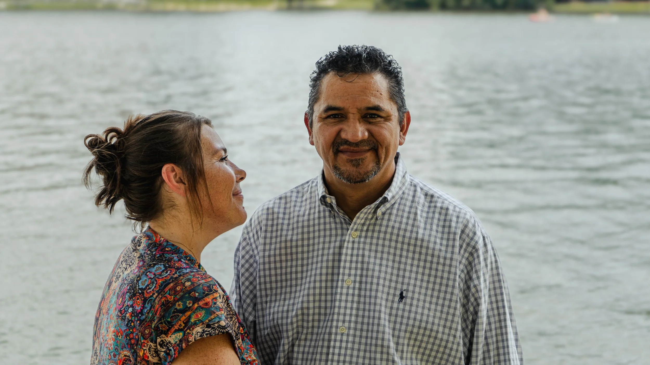 A woman and a man standing near a body of water, with the woman smiling and looking at the man, and the man smiling directly at the camera. The woman has her hair in a bun and is wearing a colorful patterned dress, while the man has dark curly hair a