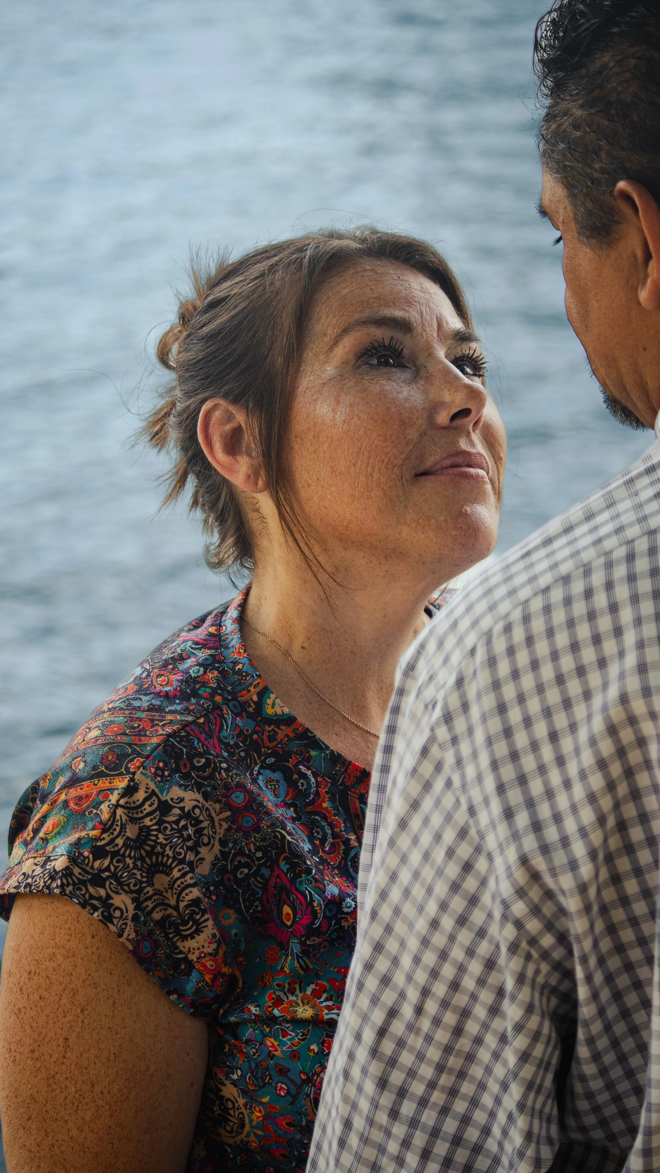 A woman with tousled brown hair, wearing a colorful patterned top, looks up at a man with dark hair, wearing a checkered shirt, with a body of water in the background.