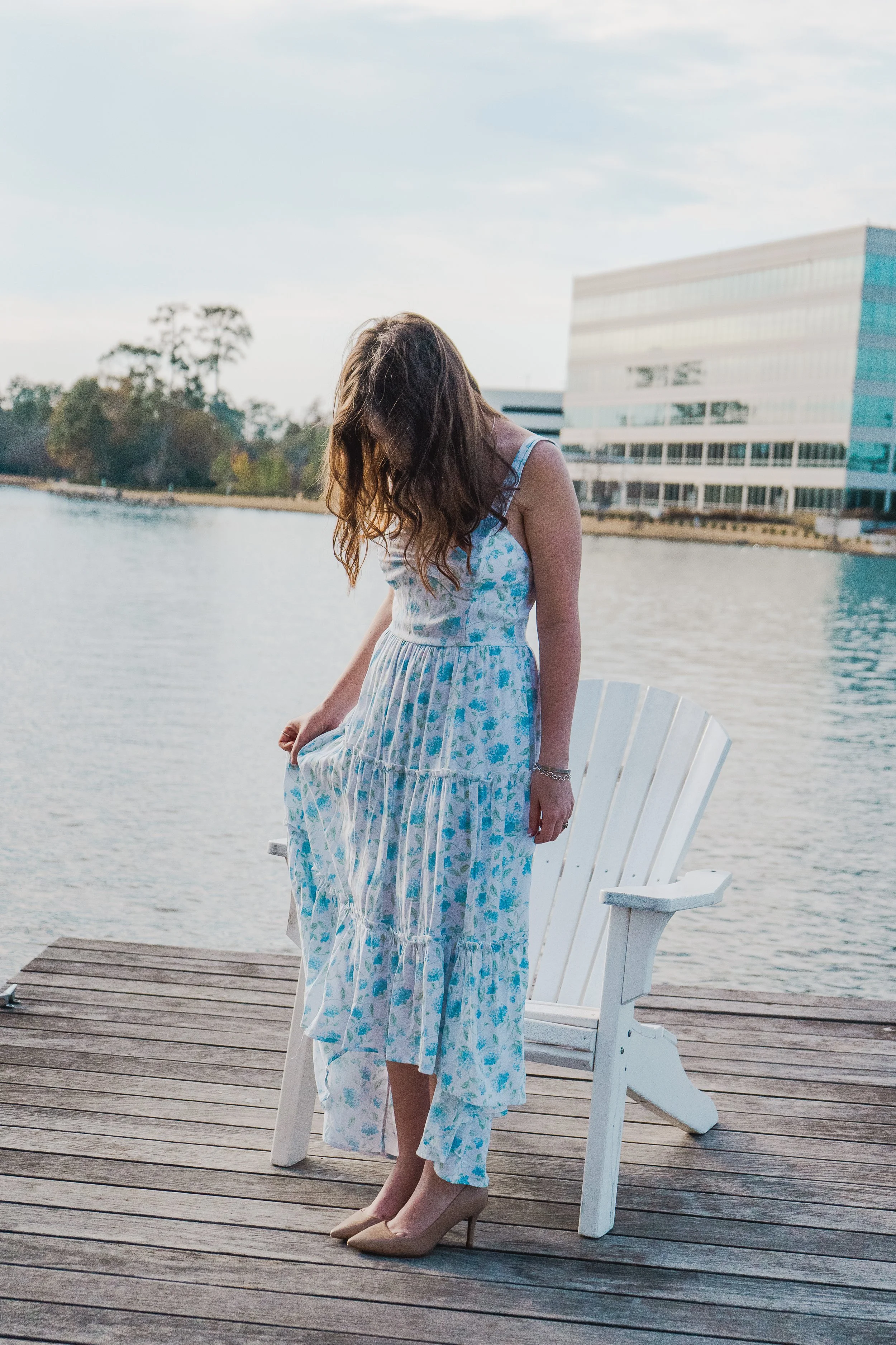 A woman in a long, flowing blue and white floral dress and nude high heels standing on a wooden dock by the water, with her head bowed and hair covering her face. There is a white Adirondack chair beside her and modern buildings in the background.