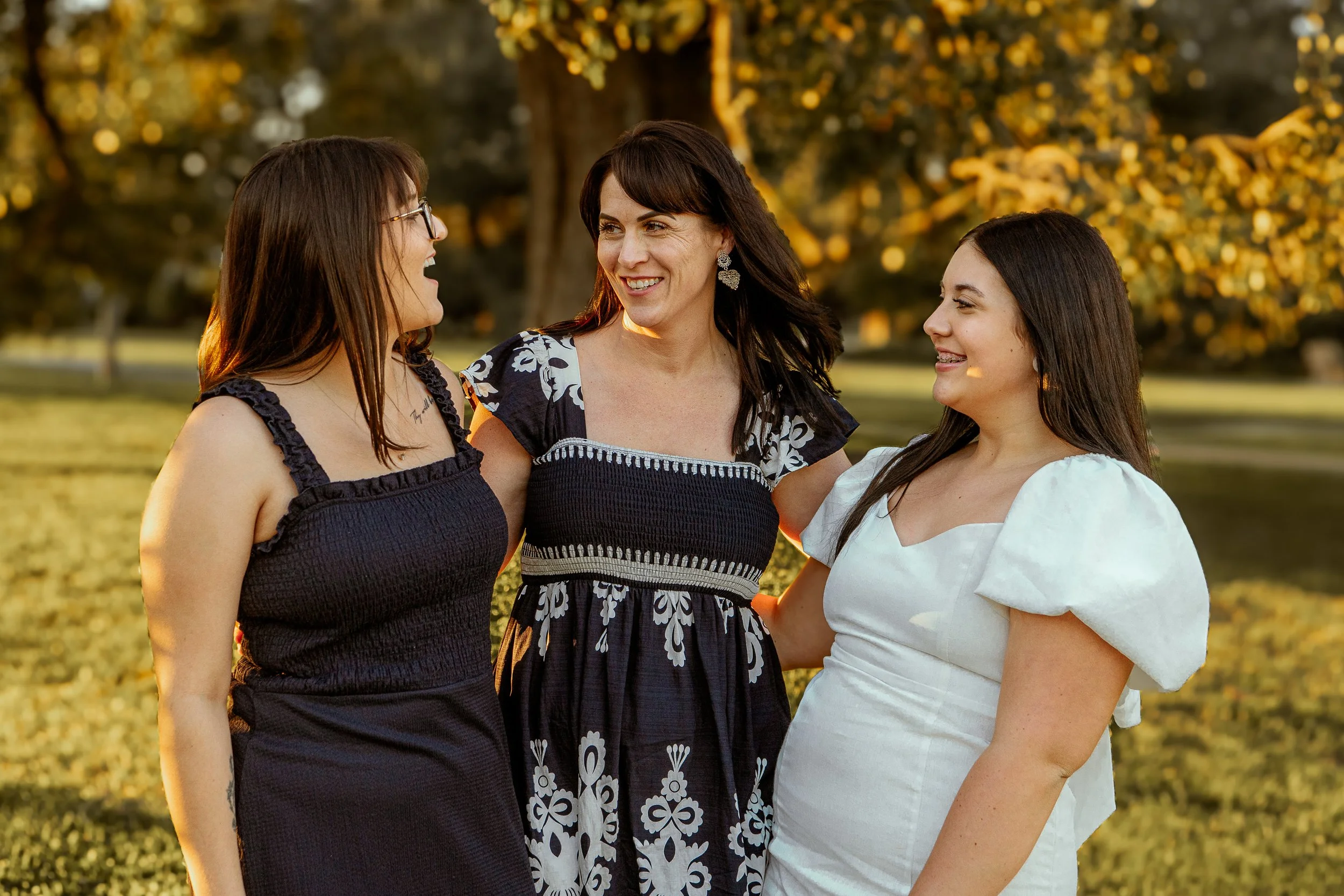 Three women standing outdoors in a park during golden hour, engaging in conversation and smiling at each other.