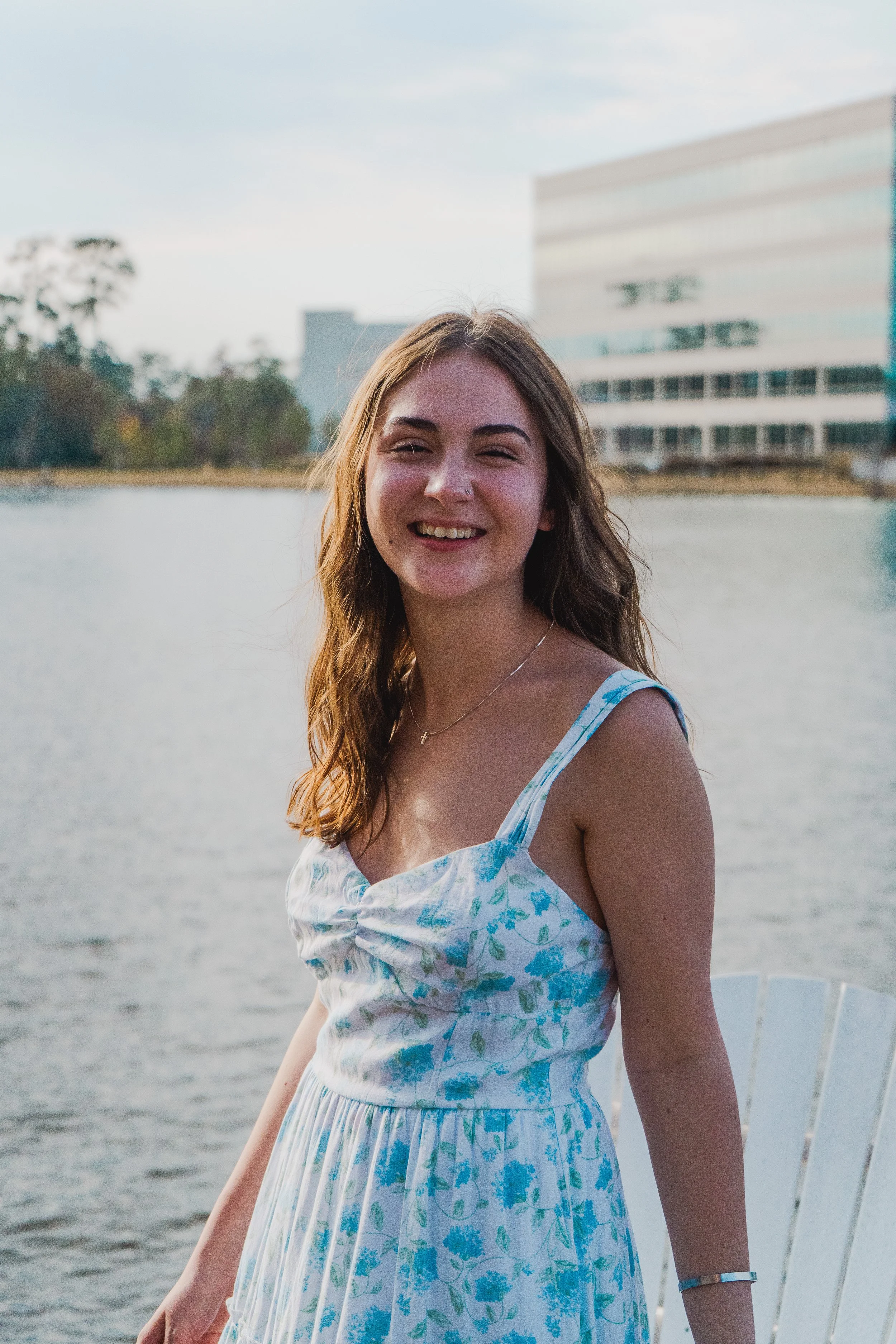 Young woman with long brown hair, wearing a blue and white floral sundress, smiling near a body of water with a modern building and trees in the background.