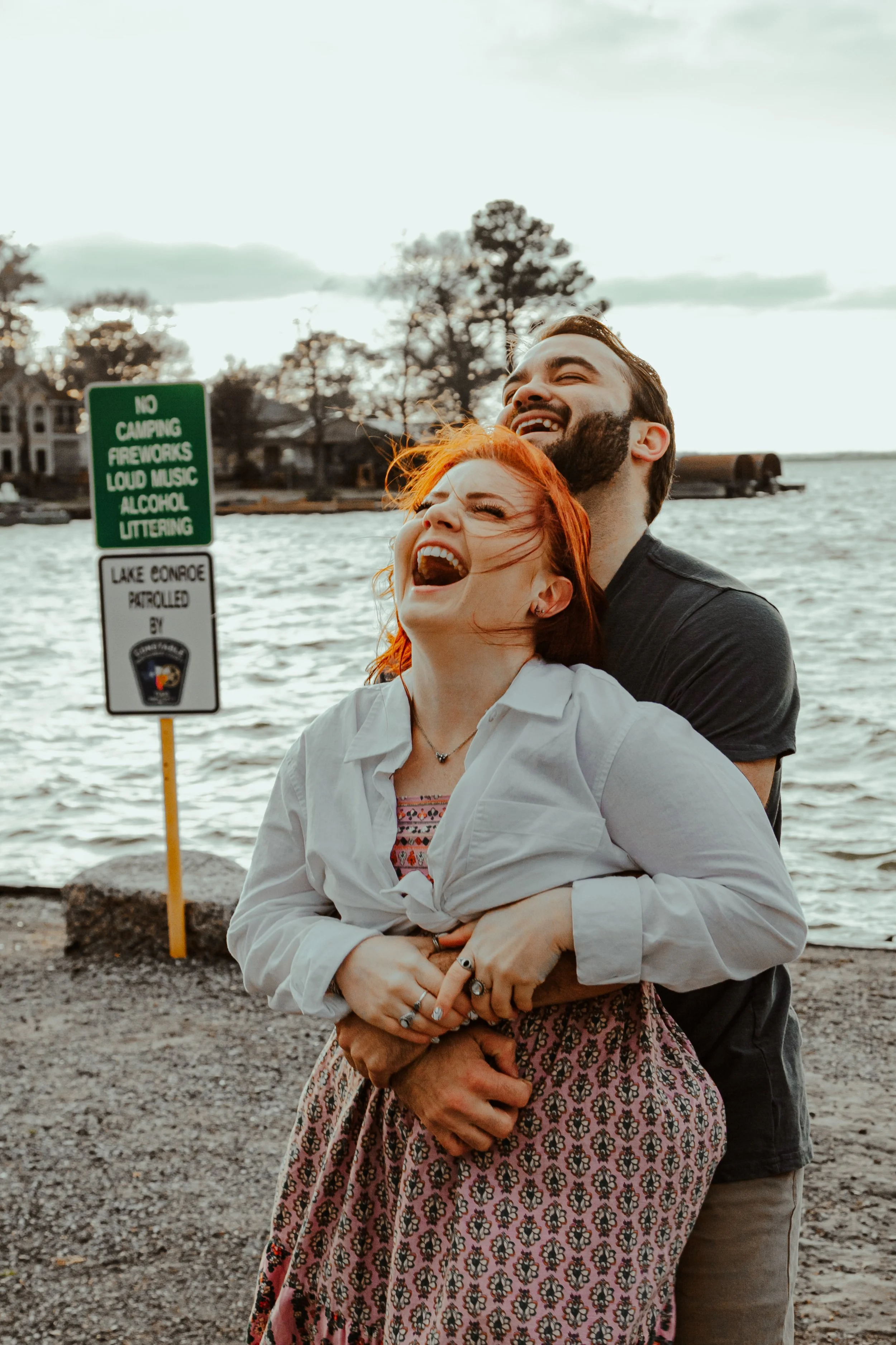 A happy couple is laughing and embracing near a lake, with trees and houses in the background, during sunset.