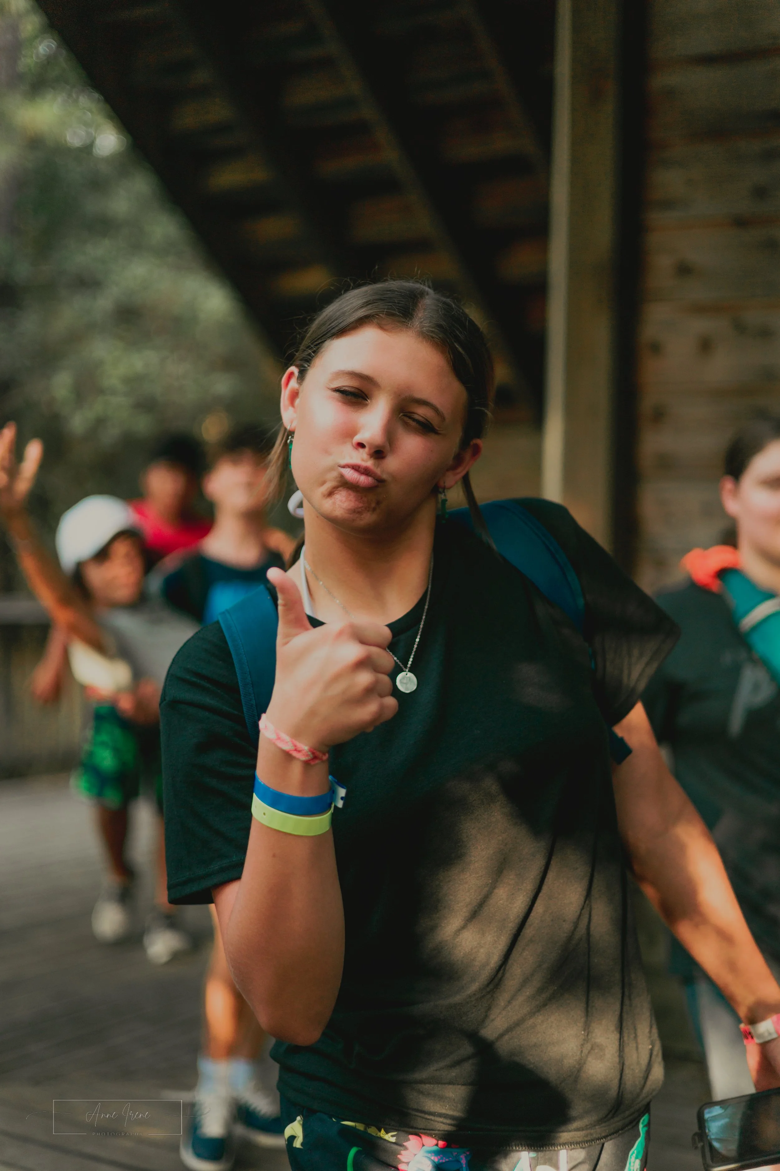 A young woman with dark hair making a duck face and giving a thumbs up while standing outdoors with a group of people in the background.