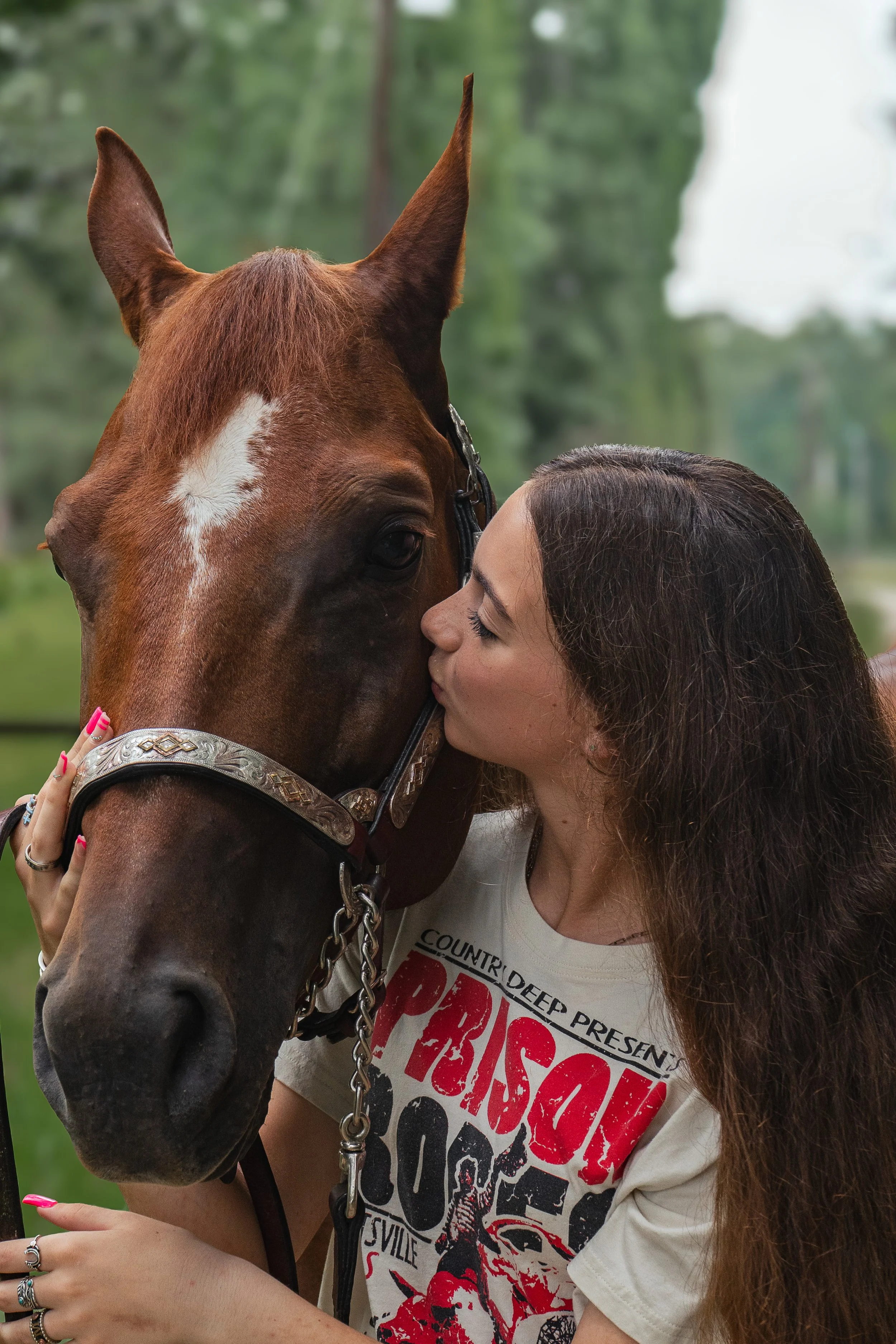 A woman with long brown hair and a white graphic T-shirt embraces a brown horse with a white star on its forehead in a green outdoor setting.