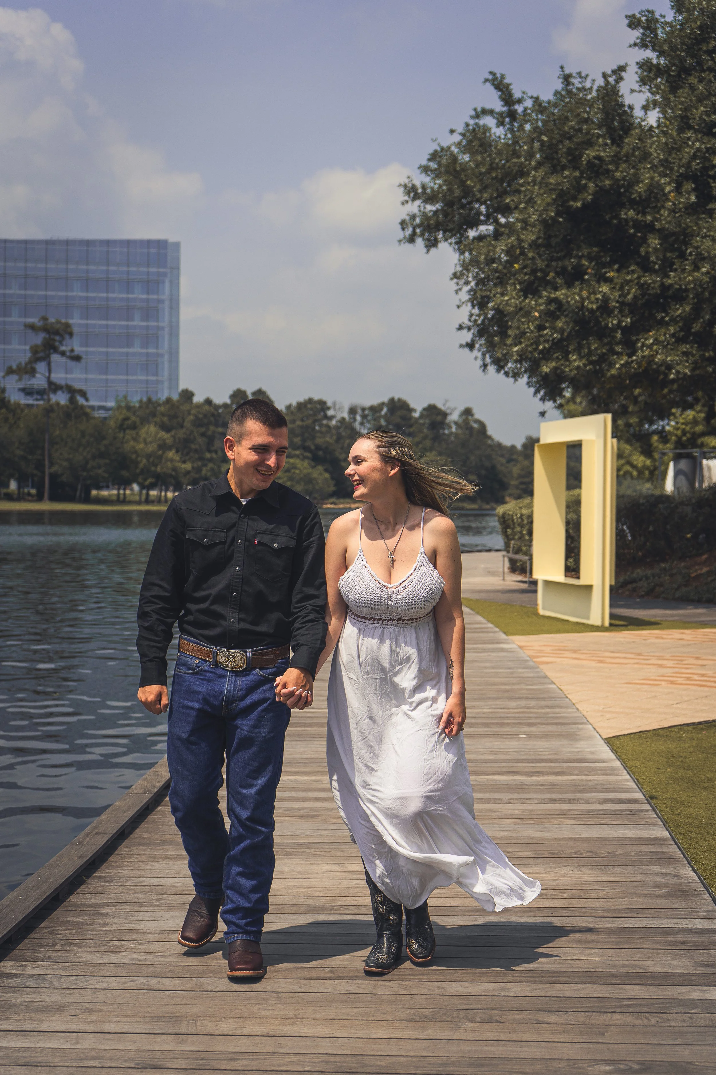 A young couple walking hand-in-hand on a wooden dock by a body of water, smiling and looking at each other on a sunny day with trees and a modern building in the background.