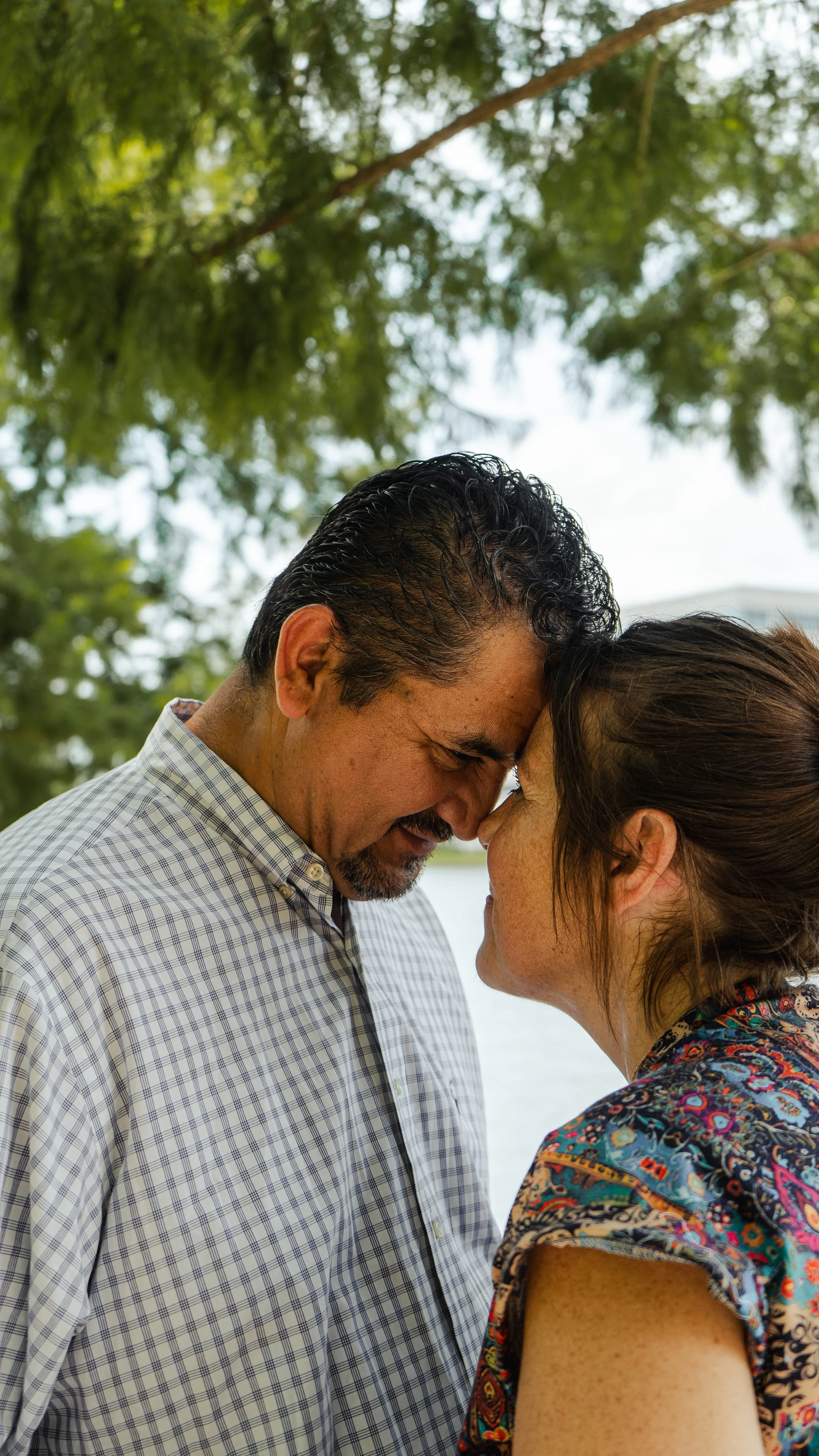 A man and woman are touching foreheads with eyes closed outdoors in a close, intimate moment, with trees in the background.