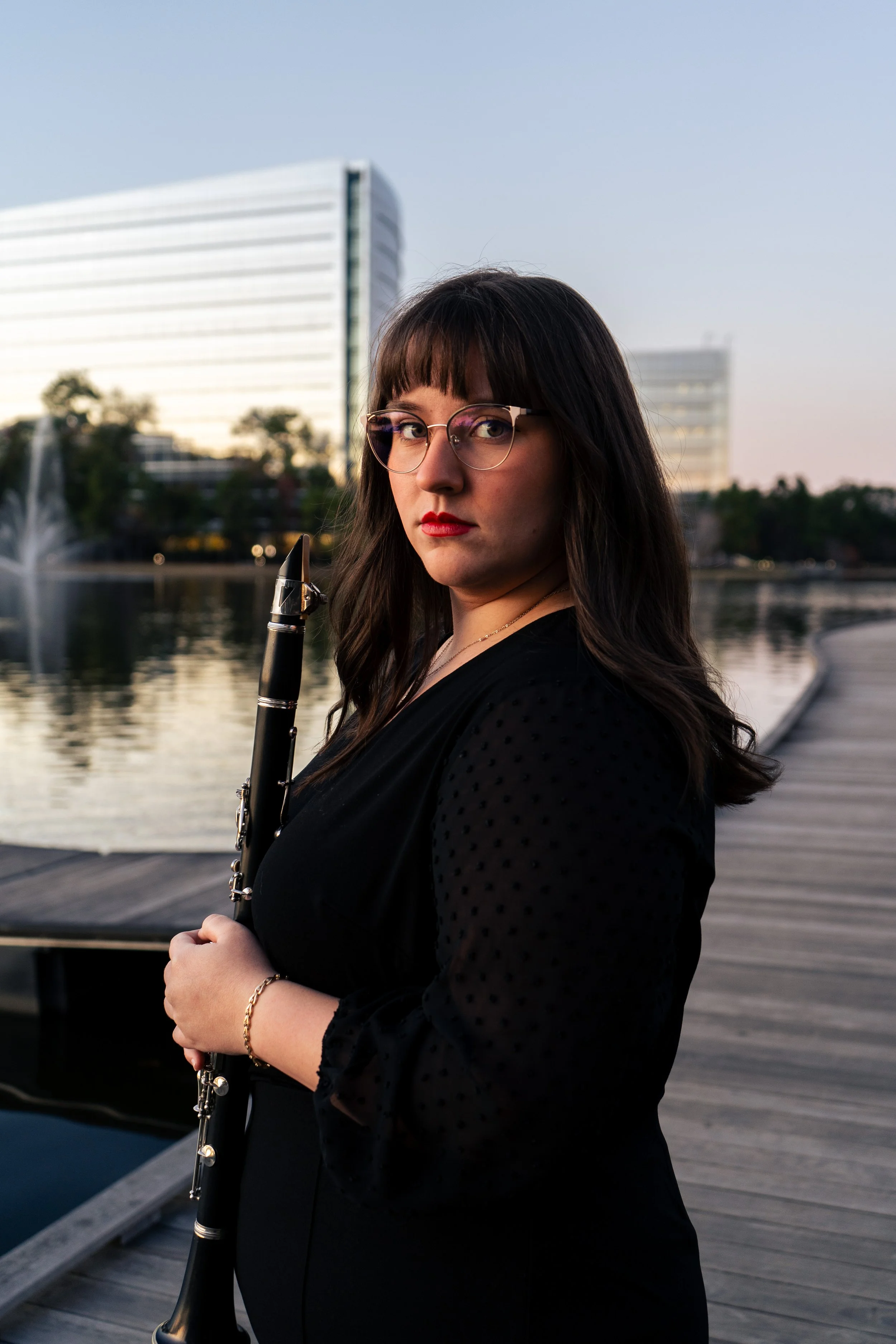 A woman with dark hair and glasses holding a clarinet, standing on a wooden dock near a body of water with modern buildings in the background during sunset.