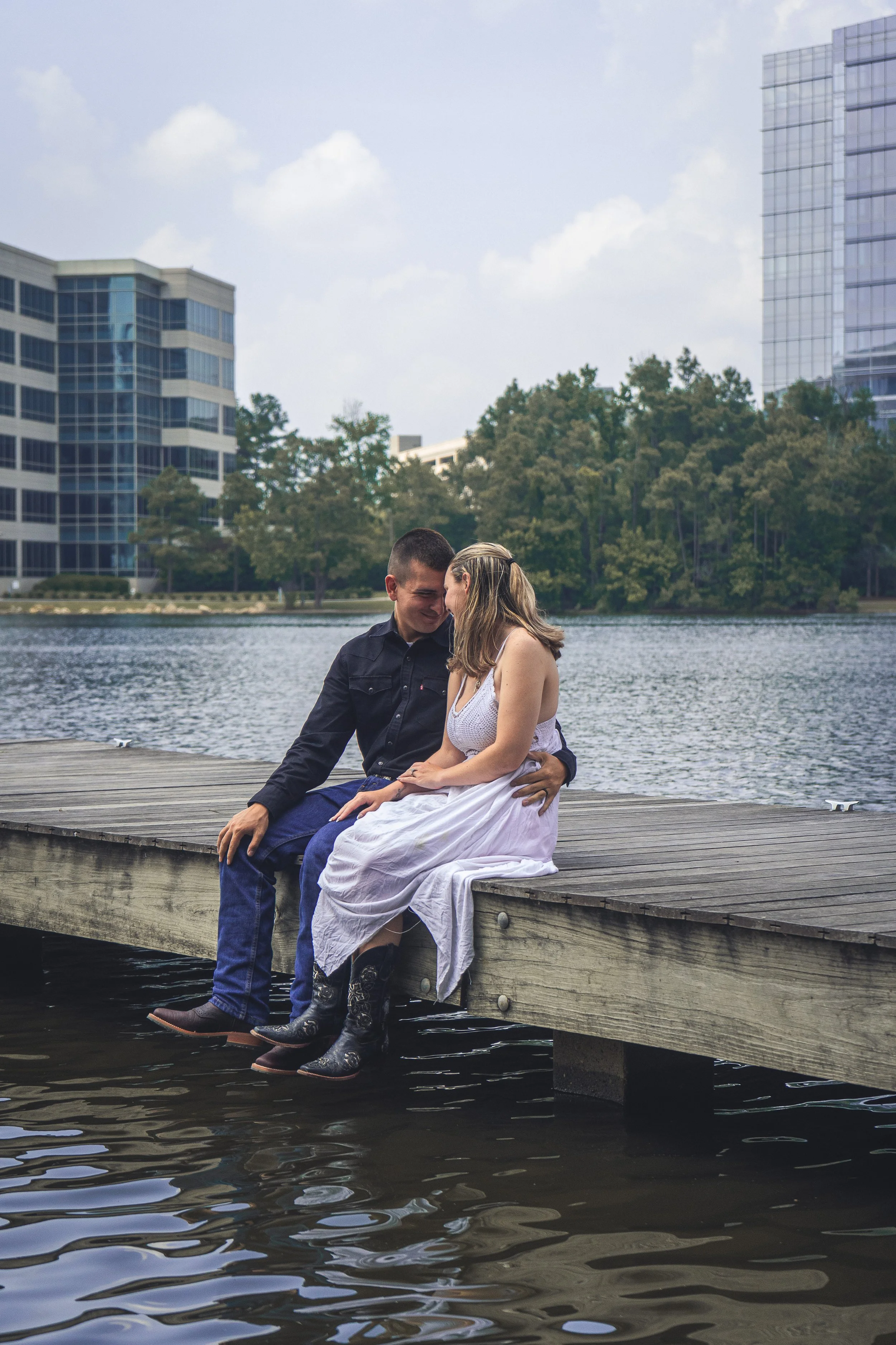 A couple sitting on a wooden dock by the water, sharing an intimate moment with their foreheads touching, surrounded by urban buildings and trees in the background.