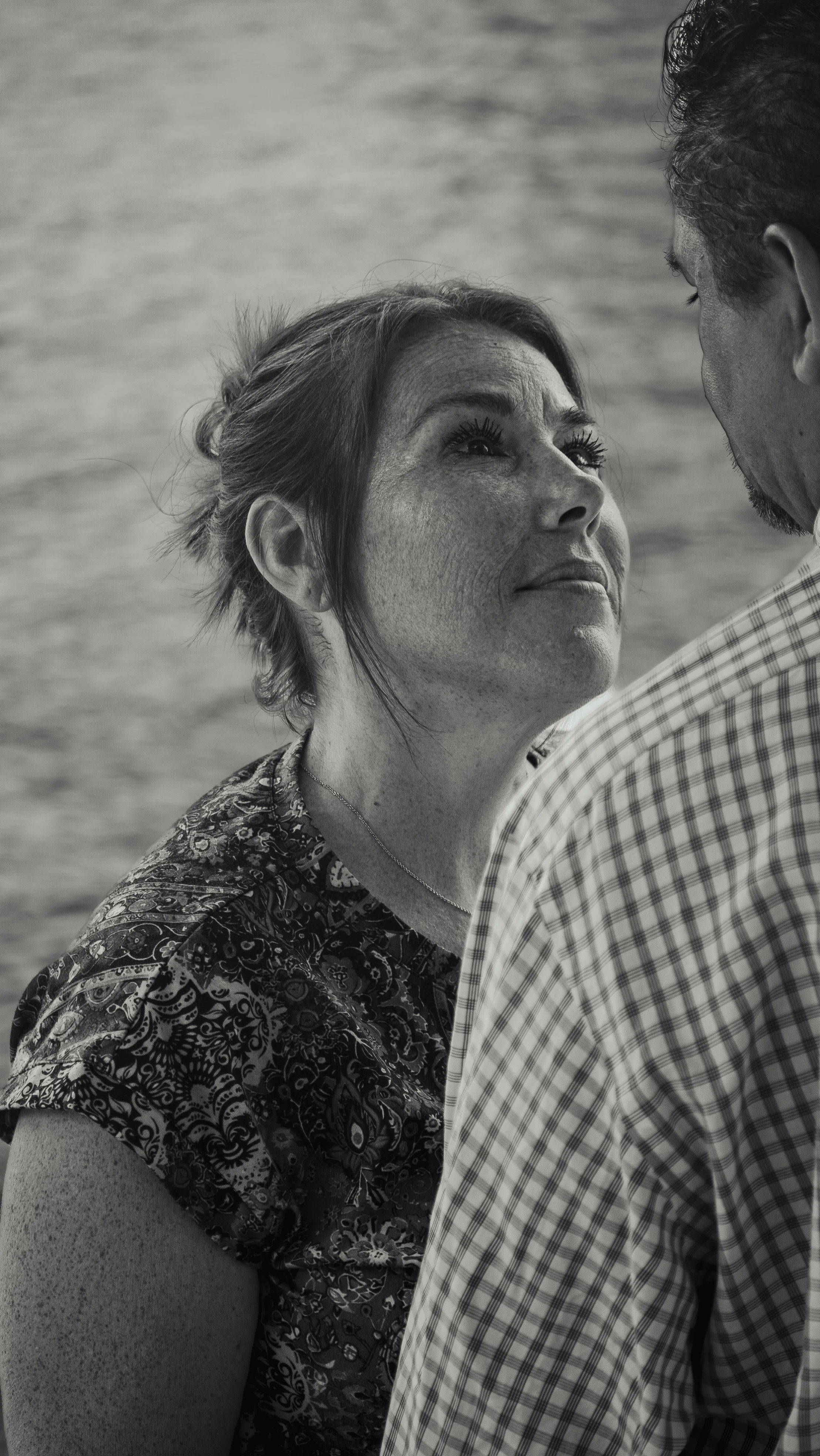 A woman looking up at a man, with her face close to his, by the water, in black and white.