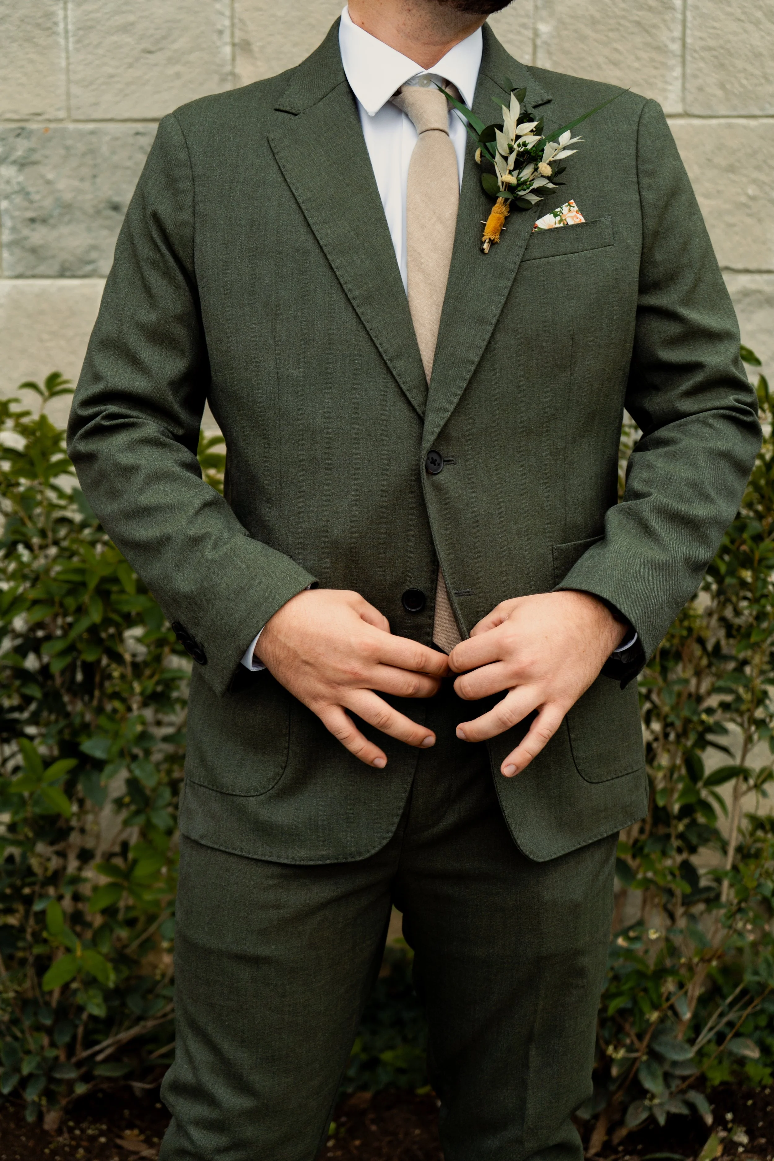 Close-up of an individual dressed in a green suit, white shirt, and beige tie, standing outdoors against a stone wall, with hands clasped in front and a boutonnière of greenery and flowers pinned to the lapel.