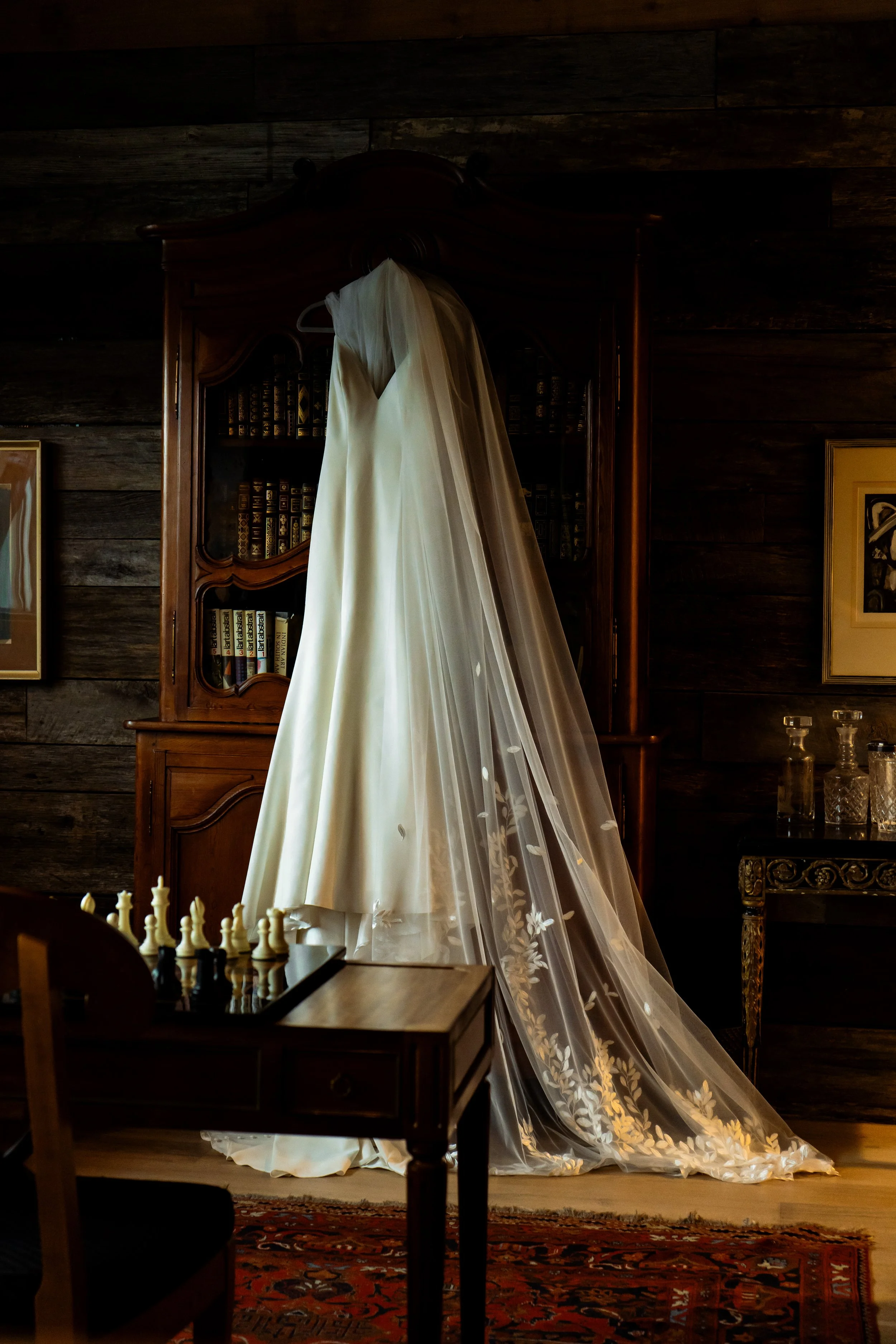 Wedding dress hanging on a wooden cabinet in a dimly lit room with dark wooden walls and a chessboard on a table in the foreground.
