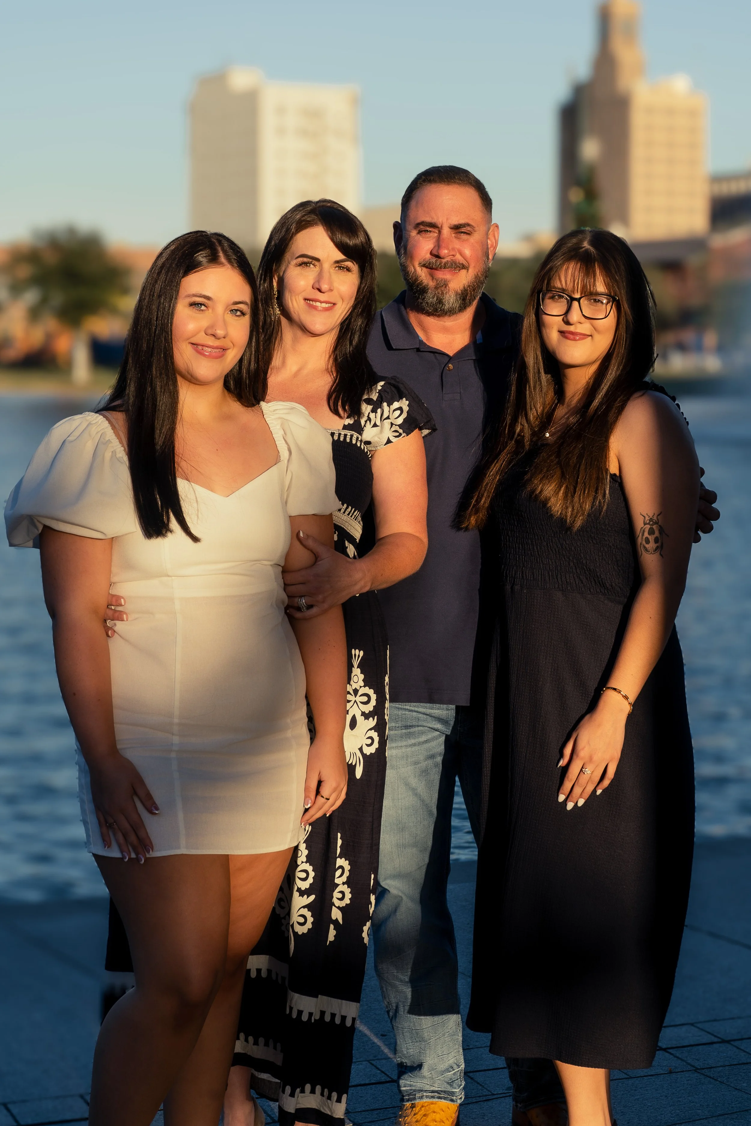 Four people, three women and one man, standing close together outdoors near water, with tall buildings in the background. They are smiling and posing for the photo, likely during sunset.