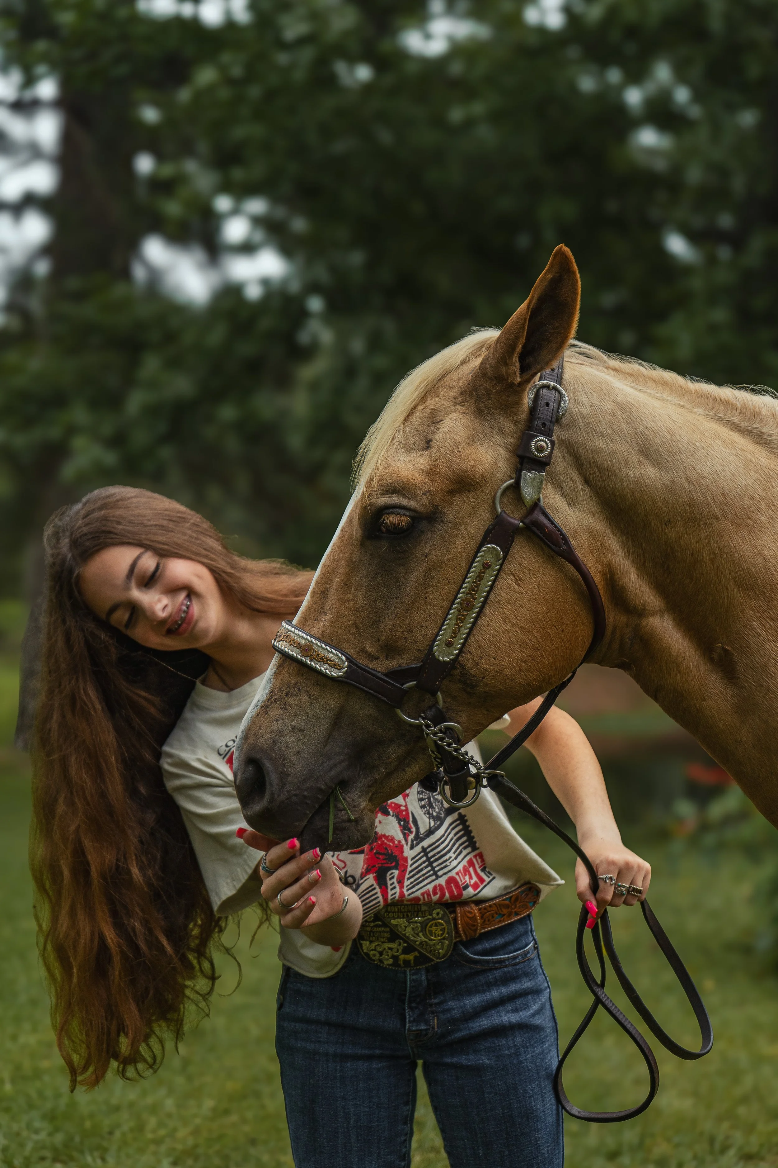 A young woman with long, wavy brown hair is smiling as she interacts with a tan horse outdoors on a grassy area. The horse is wearing a decorative bridle and is eating grass, with a background of green trees.