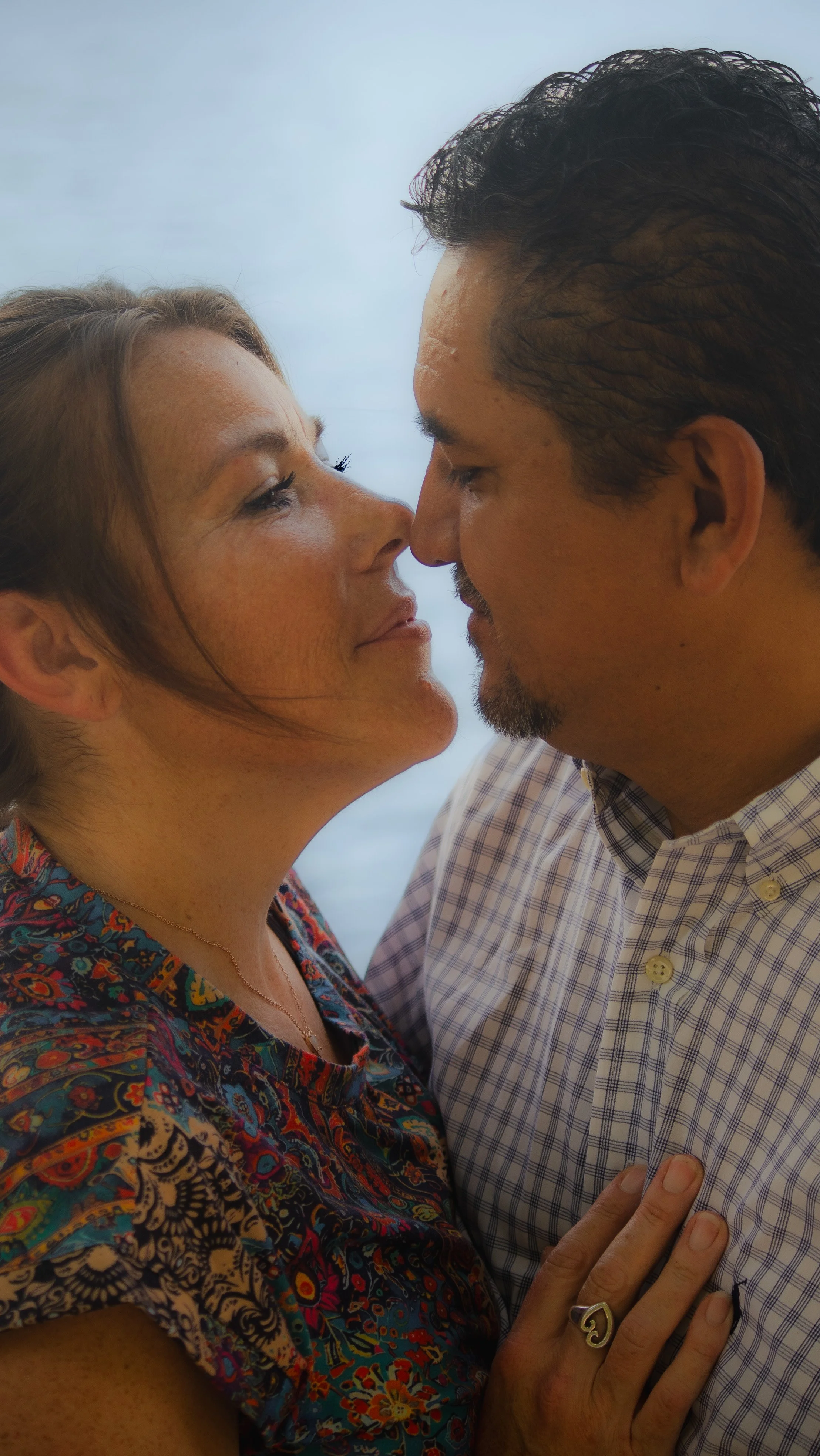 A man and a woman face each other closely with eyes closed, about to kiss, with their foreheads touching. The woman has light brown hair and is wearing a colorful patterned top, while the man has dark hair and is wearing a plaid shirt. The woman’s le