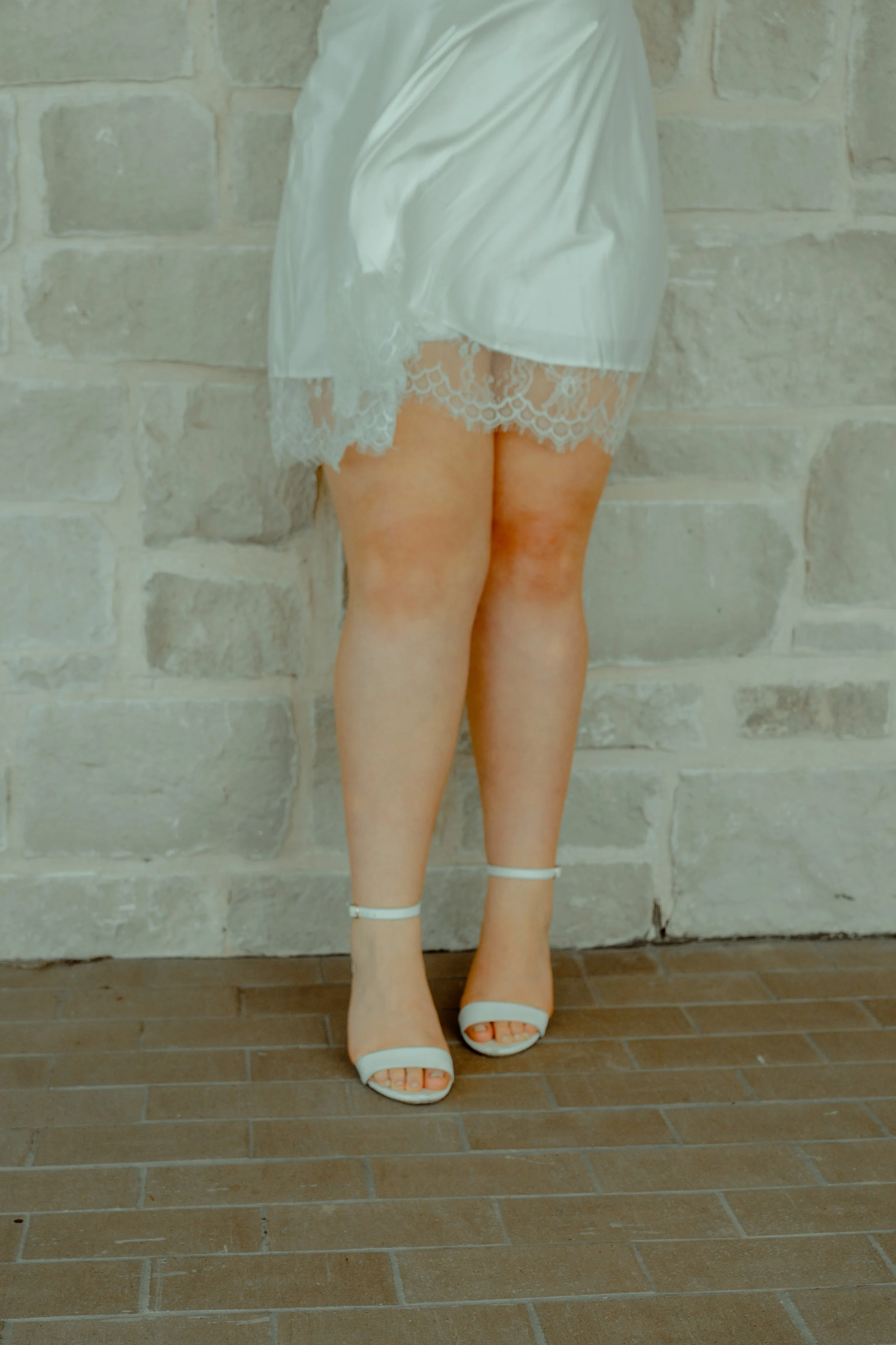 A woman standing in front of a brick wall, wearing a satin robe with lace trim and white high-heeled sandals.