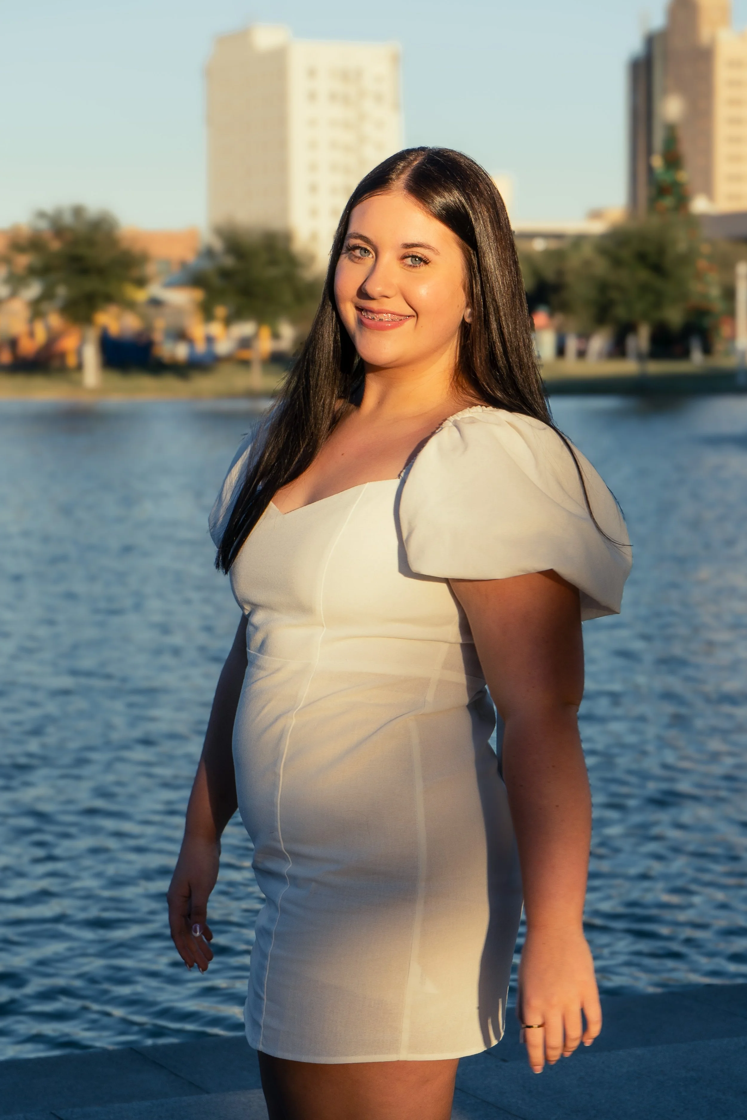 A young woman with long dark hair and a light complexion smiling, standing outdoors near a body of water with a city skyline in the background. She is wearing a light-colored dress with puffed sleeves and a ring on her right hand. The sunlight illumi