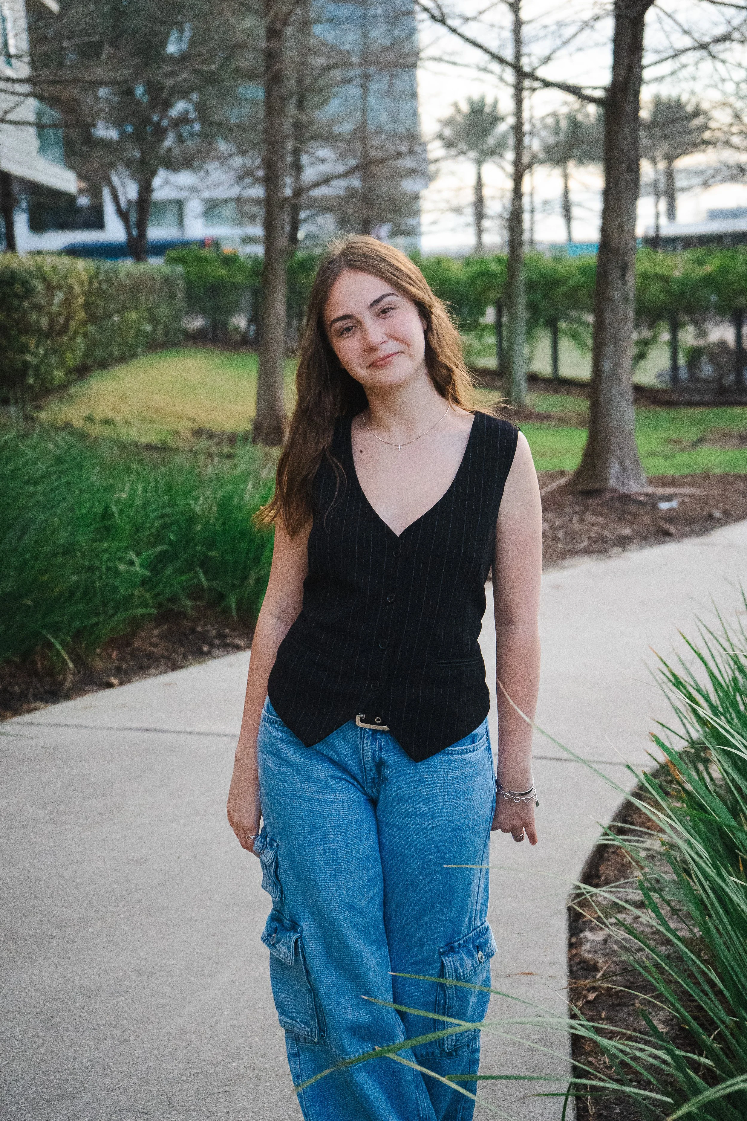 A young woman with long brown hair, wearing a black sleeveless top and blue cargo jeans, standing outdoors on a sidewalk near trees and bushes during daytime.