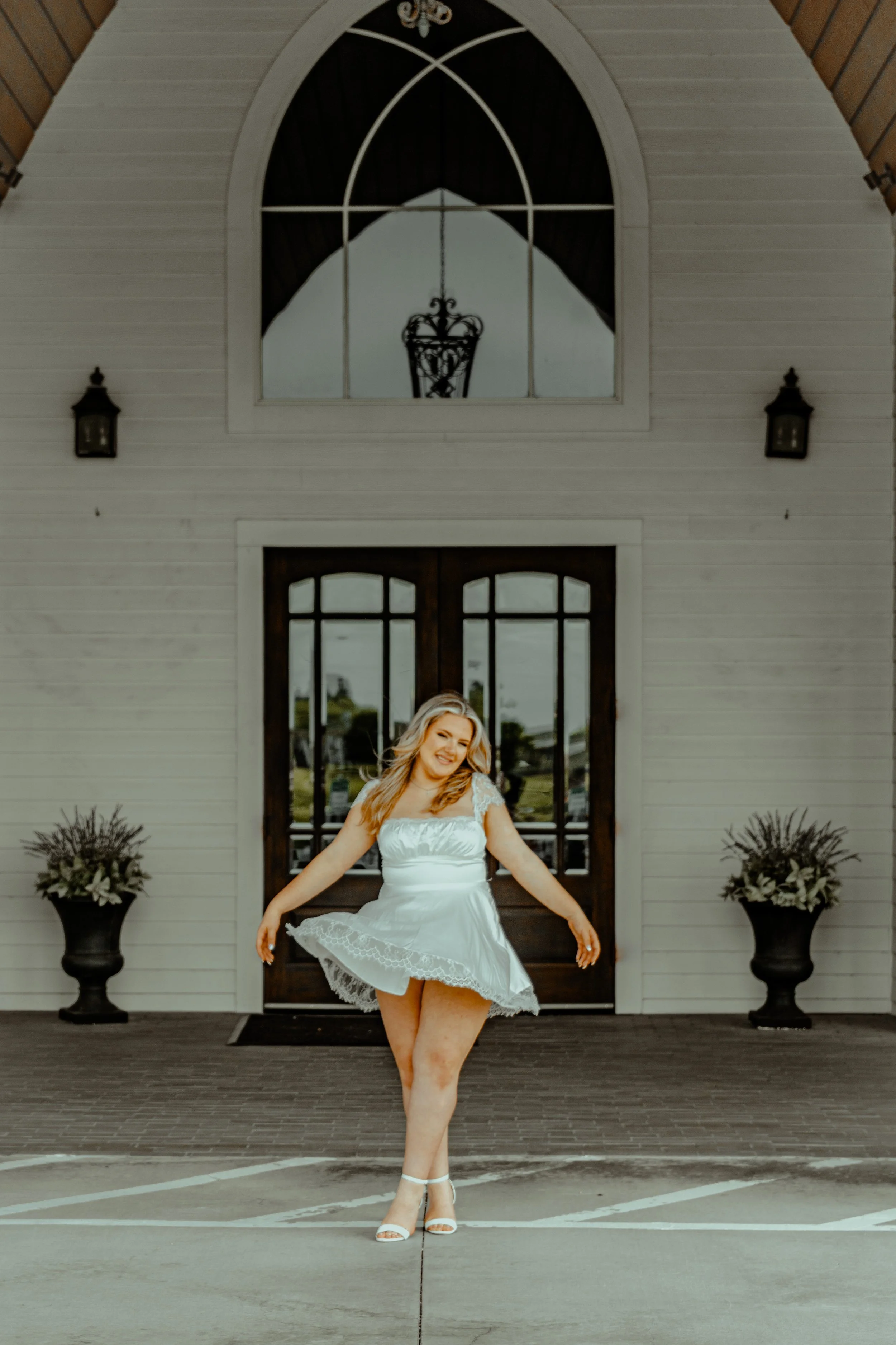 A woman in a white dress and heels standing in front of a house with a large arched window and double doors, smiling and posing with her arms outstretched.