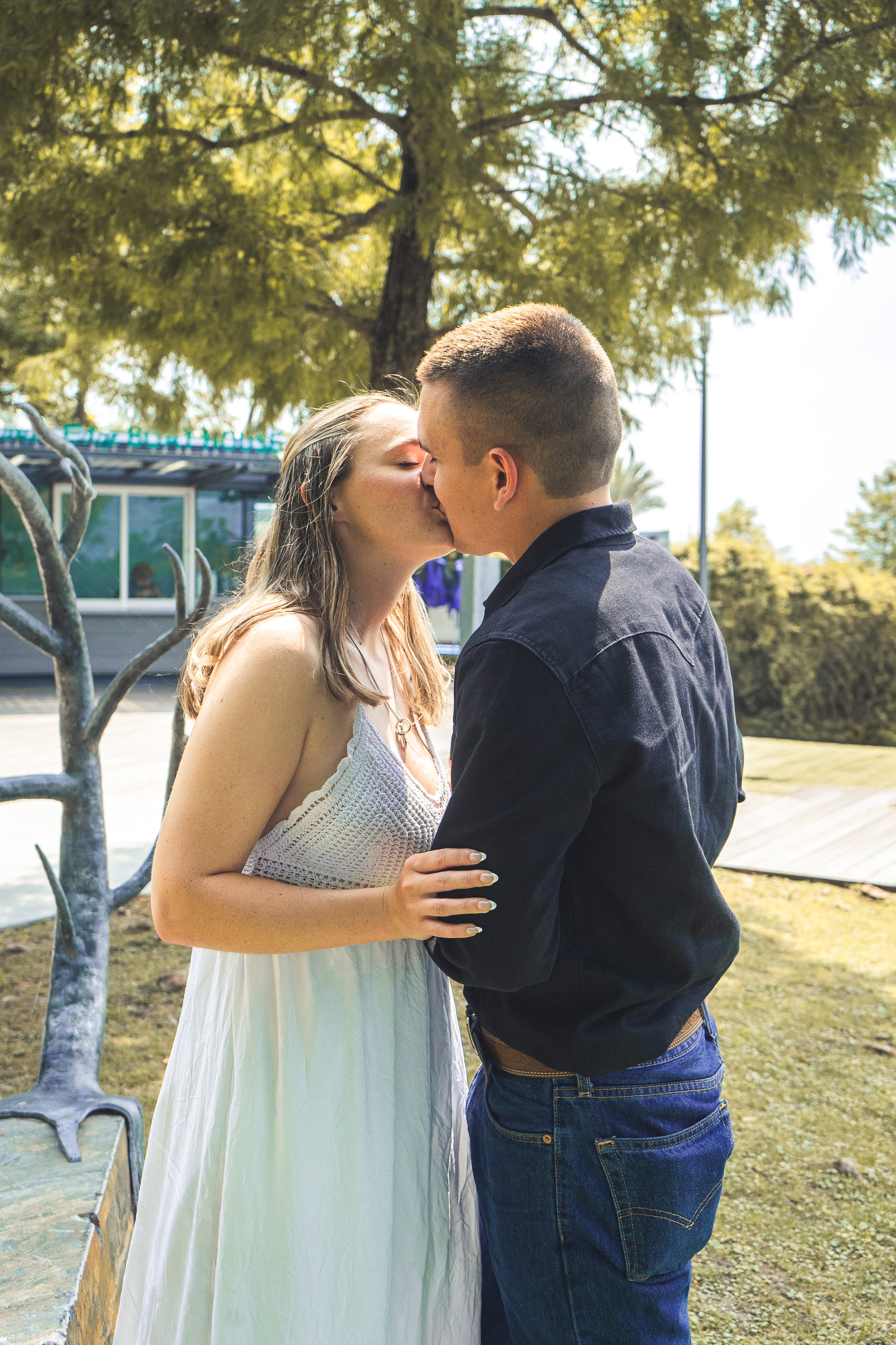A young man and woman are kissing outdoors on a sunny day. The woman is wearing a white dress, and the man is wearing a dark jacket and jeans. There are trees and a building in the background.